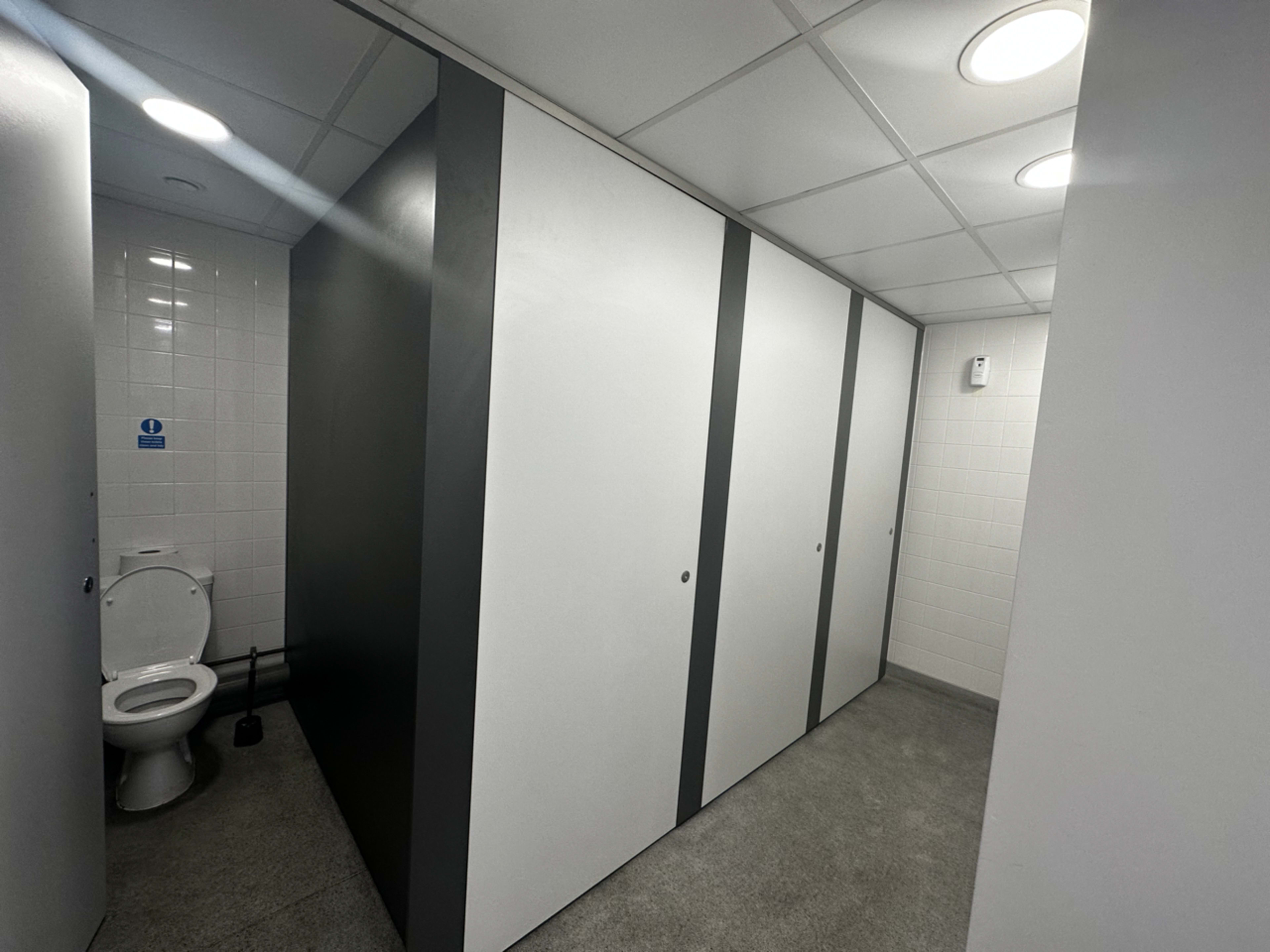 The image shows the interior of a restroom with a toilet stall on the left and multiple gray and white partitioned stalls.