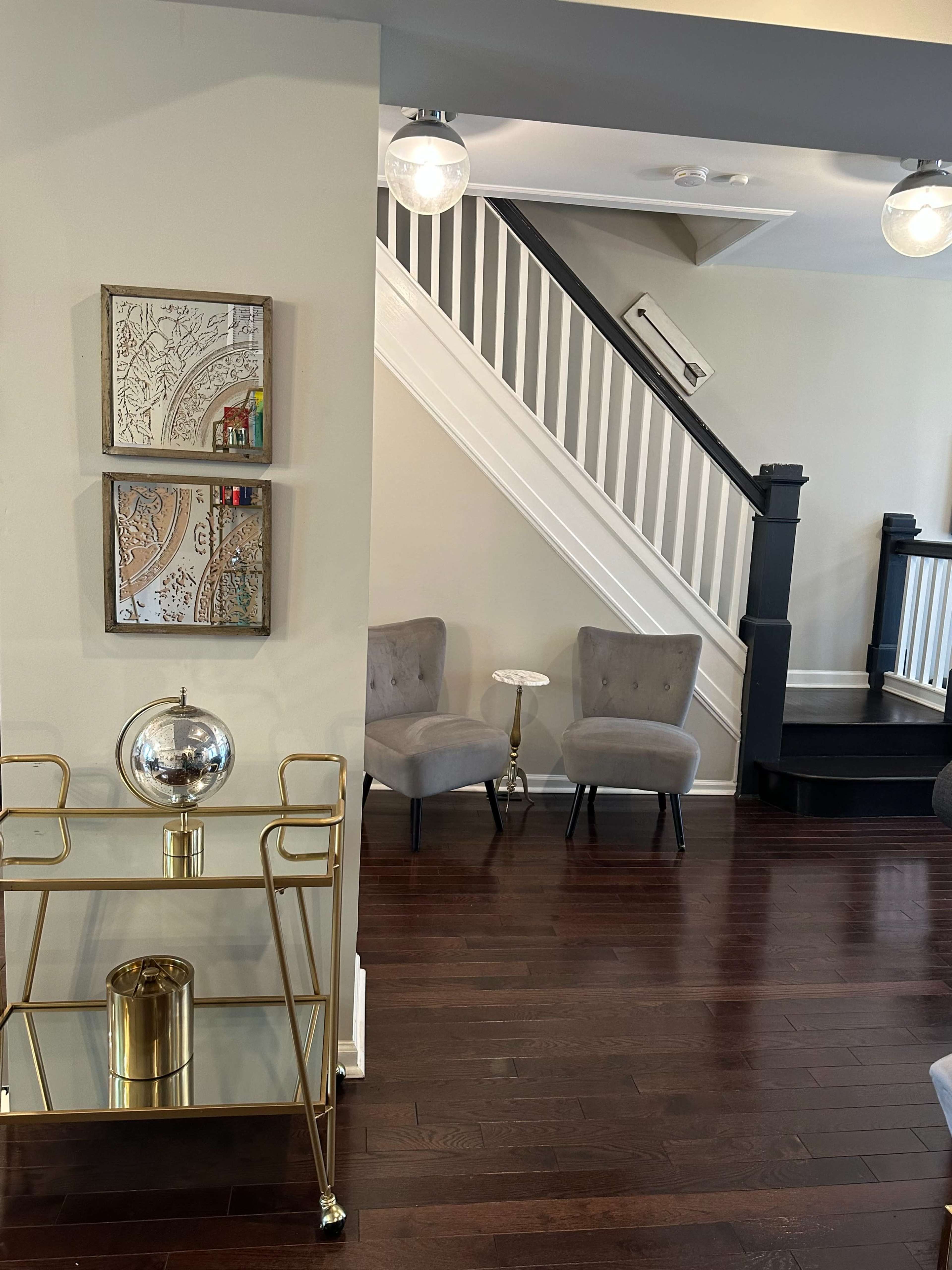 A modern interior space featuring a staircase, two upholstered chairs, and a decorative gold cart with a globe and glass container.