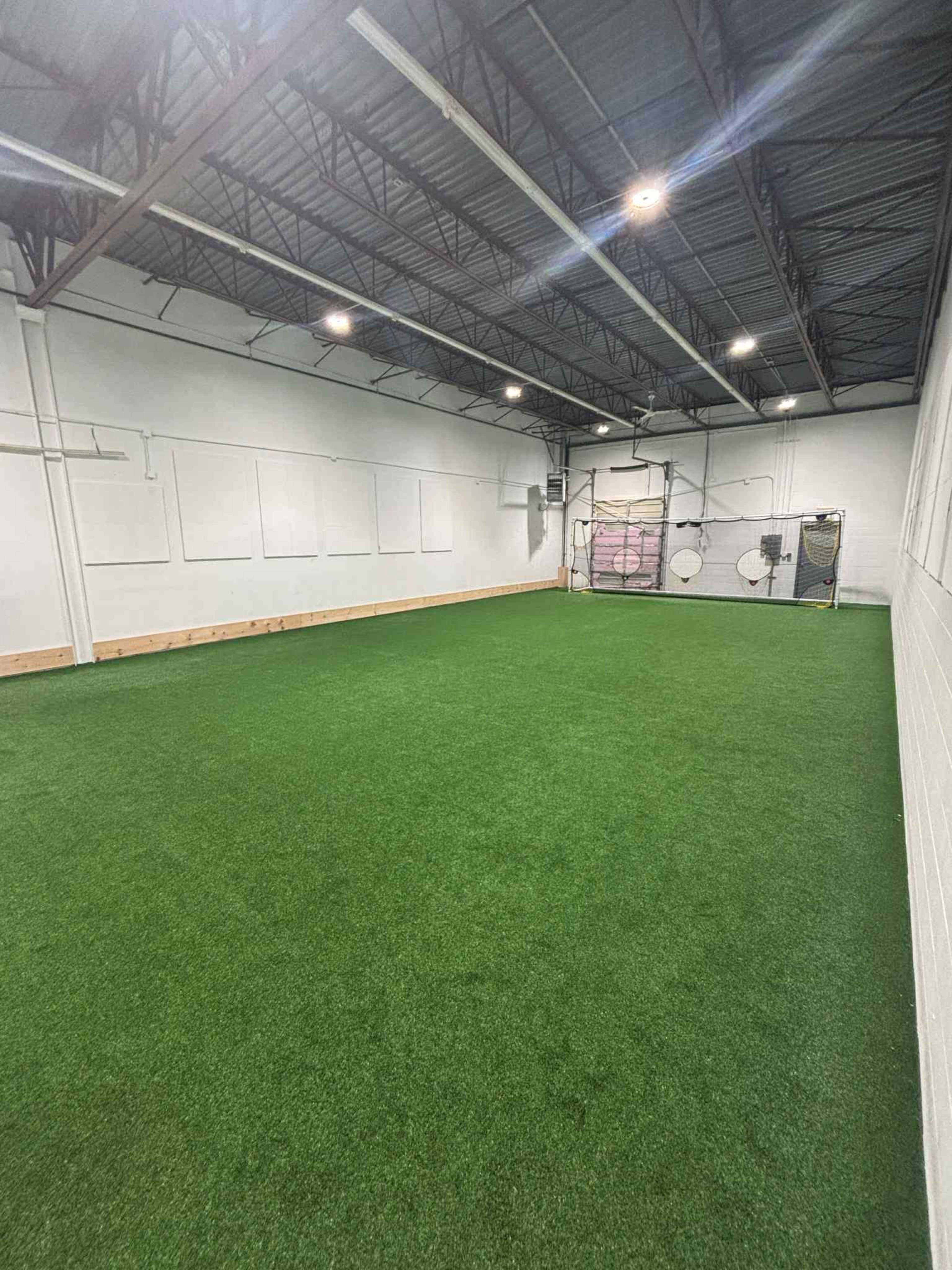 The image shows a spacious indoor soccer training facility with artificial grass and a goal at one end.