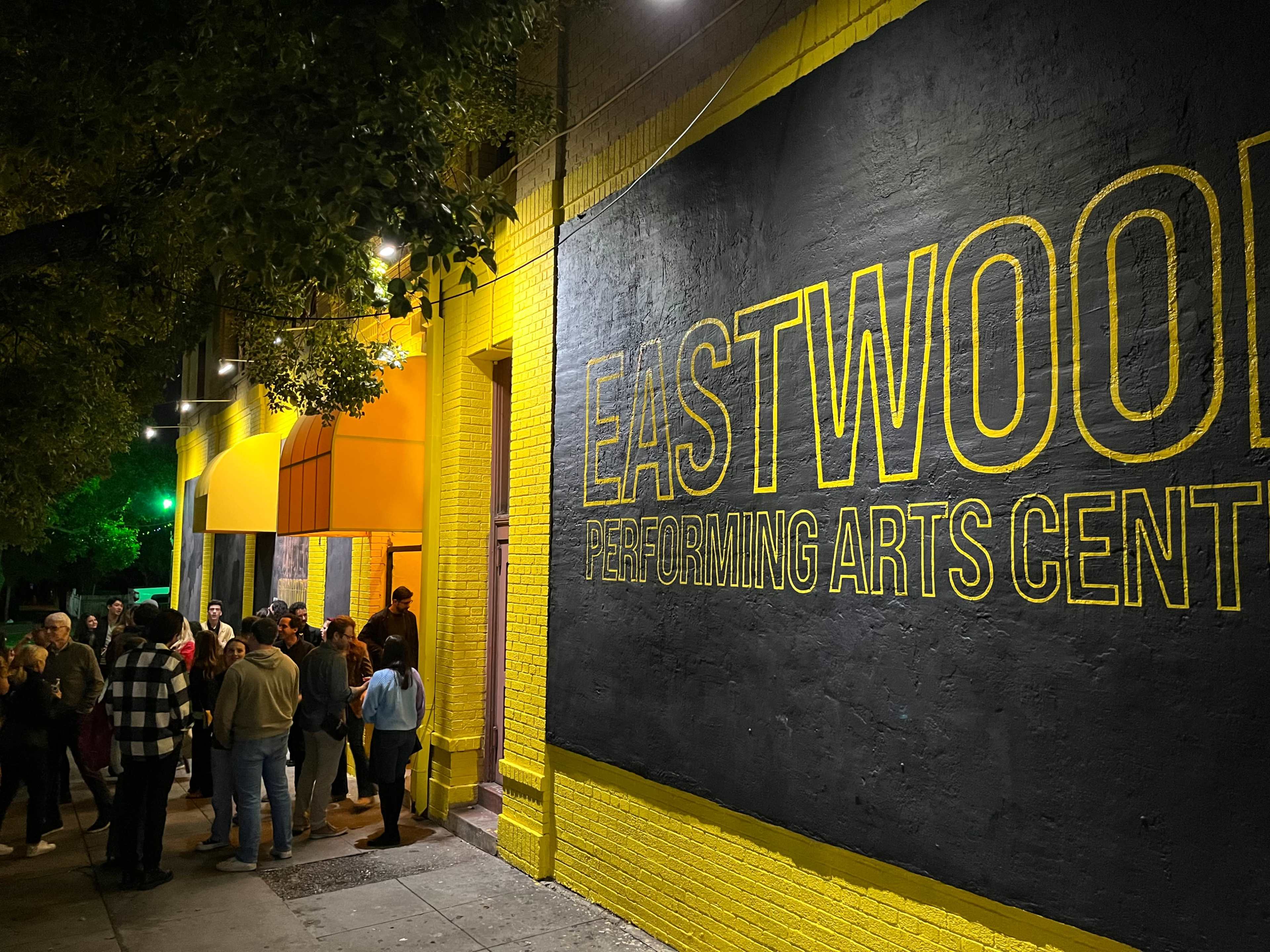 A crowd gathers outside the brightly colored Eastwood Performing Arts Center, which is illuminated by lights at night.