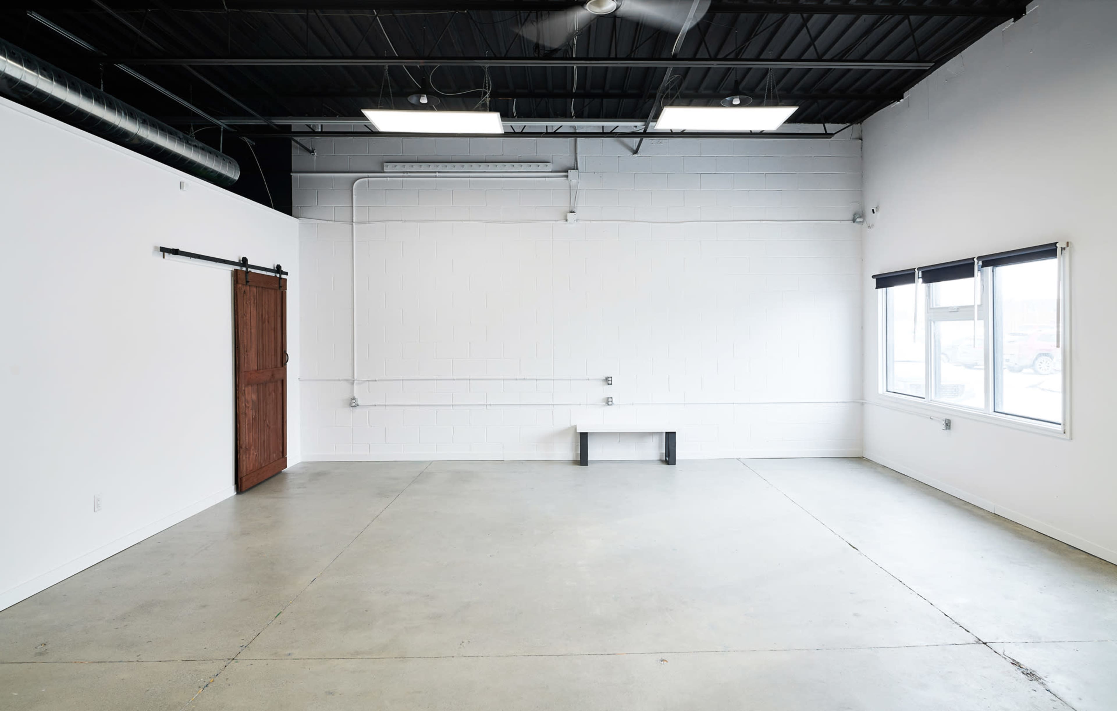 An empty room with concrete flooring, white walls, a wooden sliding door, and large windows with blinds.