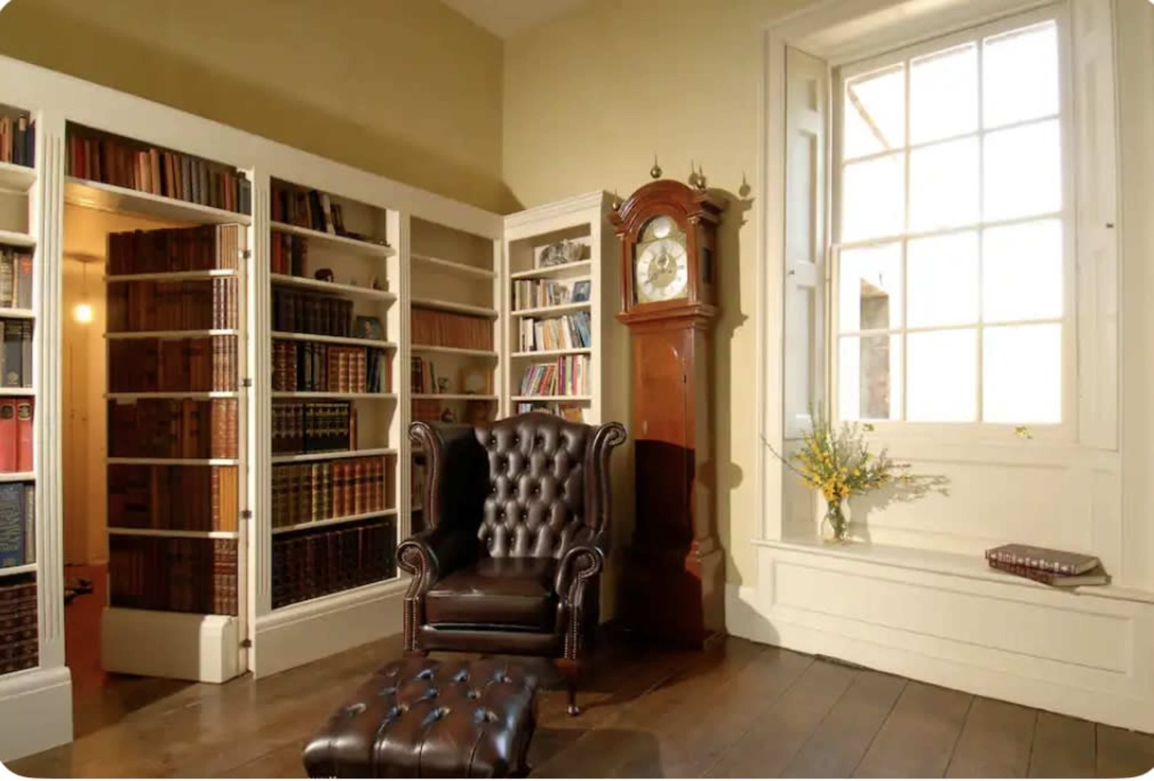 A library corner with a leather armchair, a tall grandfather clock, bookshelves filled with books, and a window allowing natural light to enter.