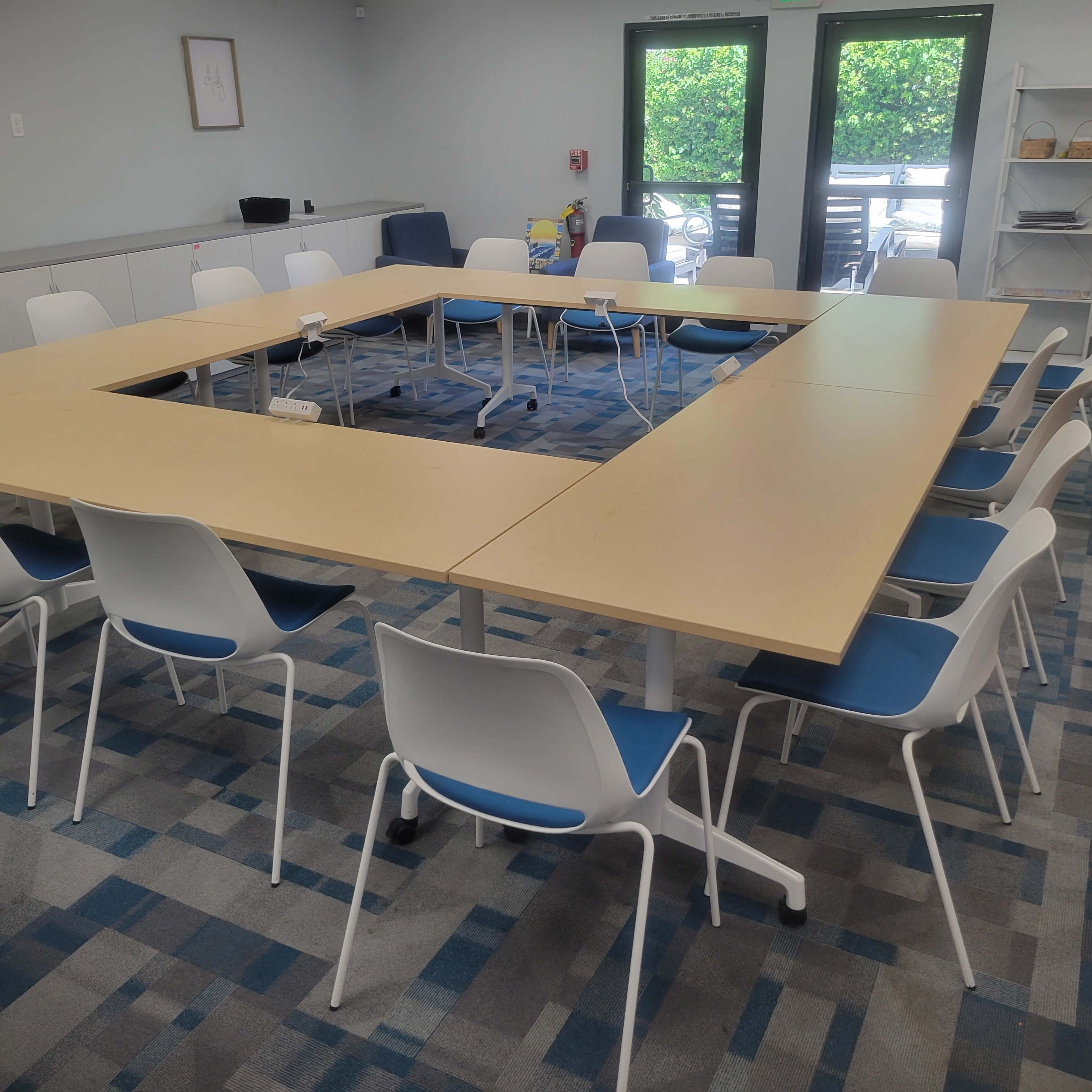 A rectangular conference table surrounded by white chairs is arranged in a meeting room with large windows and a carpeted floor.