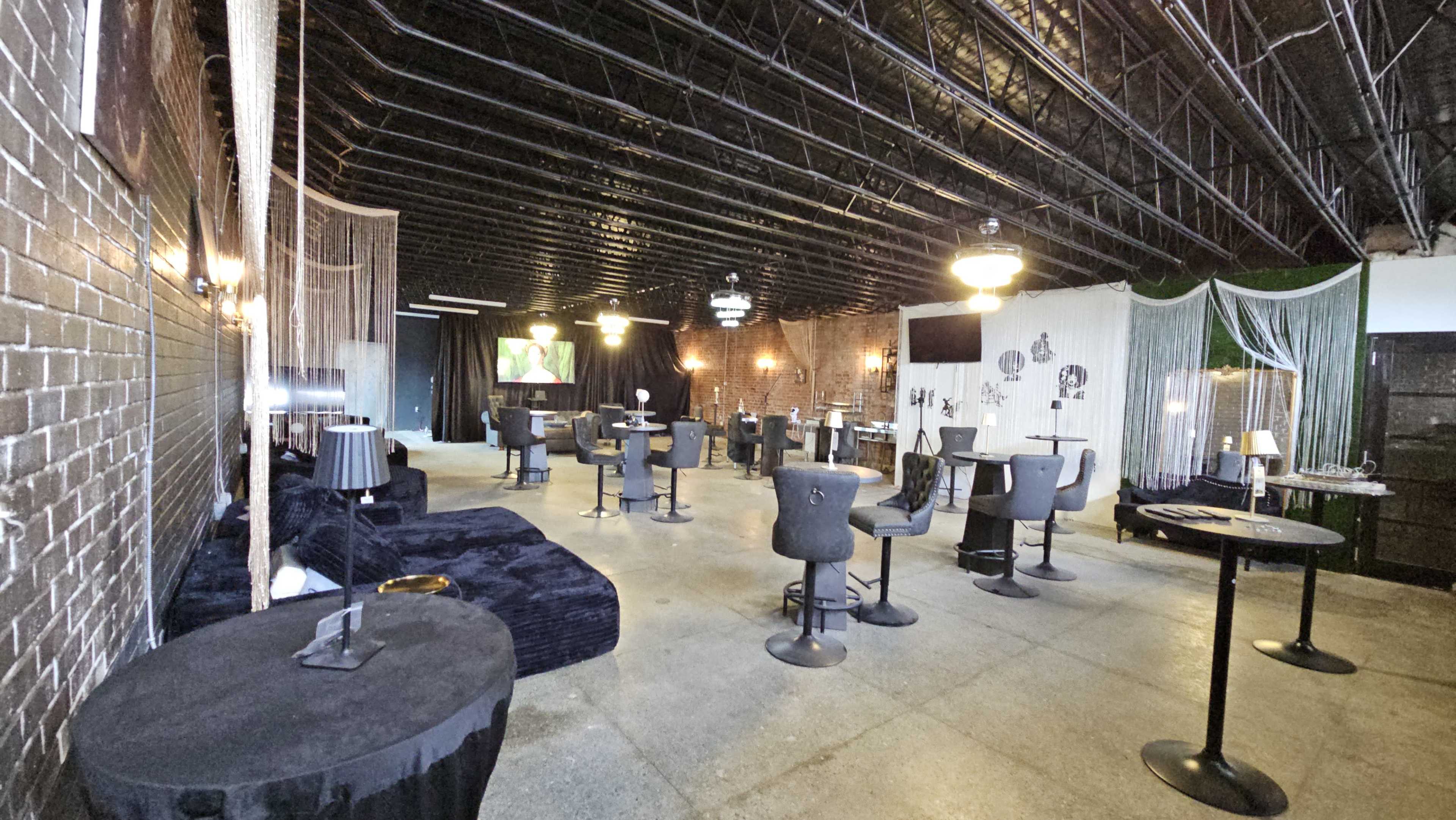 The image shows a spacious event venue with various seating arrangements, including tables and lounge areas, under a black ceiling and exposed brick walls.