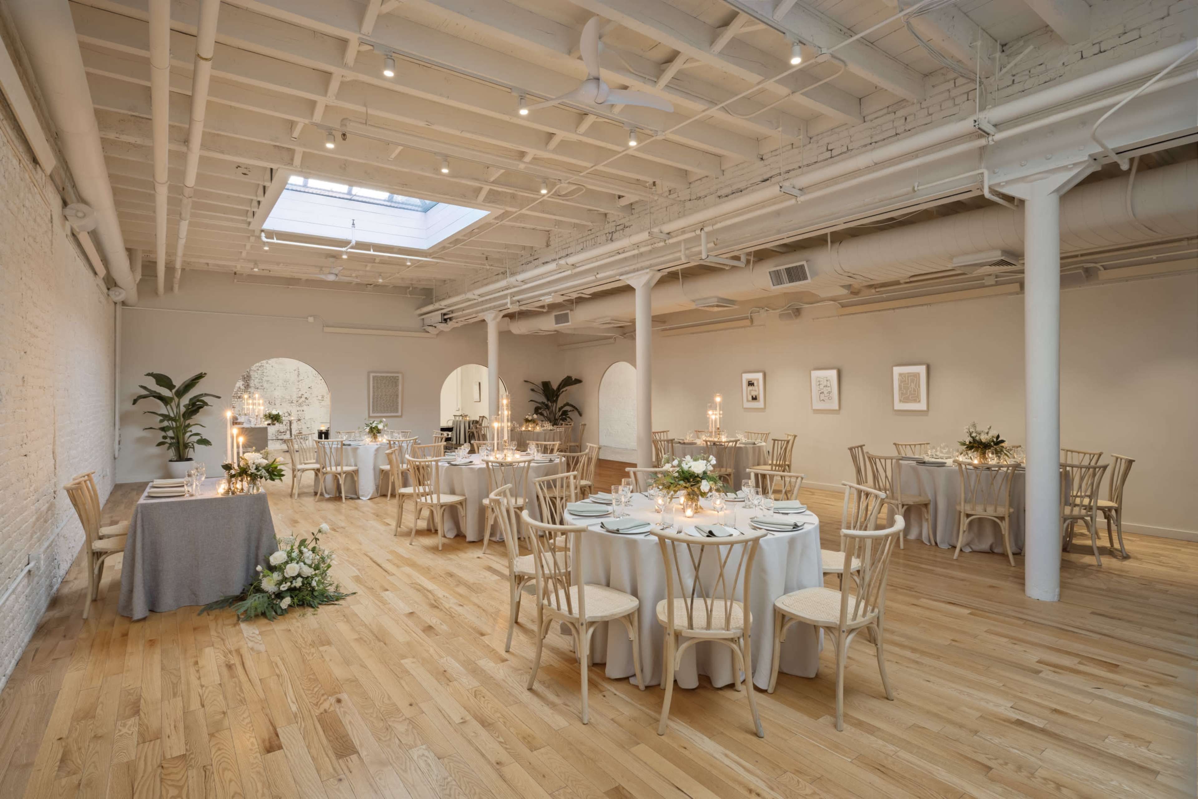 The image shows a well-lit event space with multiple circular tables set for dining, alongside elegant floral arrangements and a skylight above.
