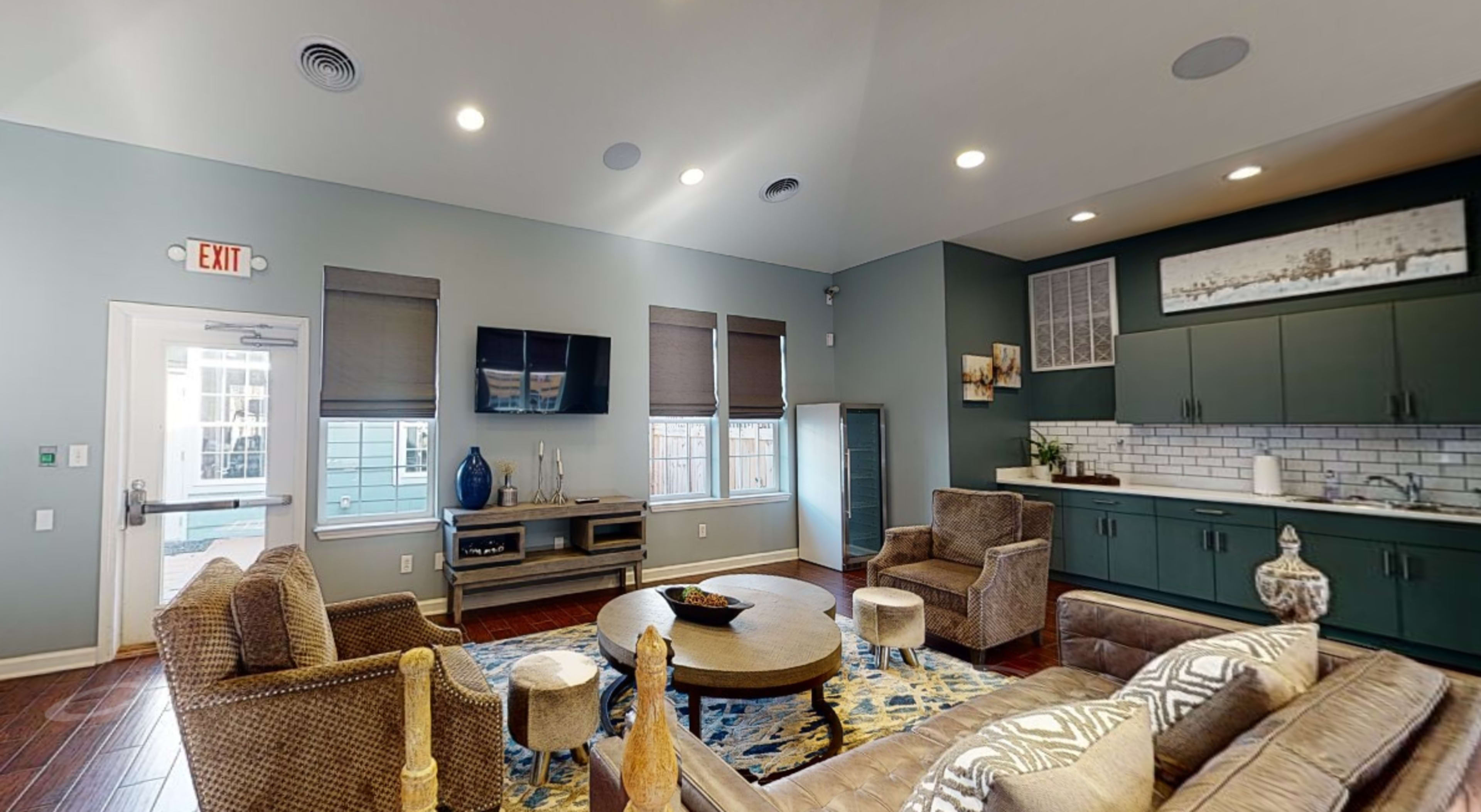 The image shows a modern living room with seating arrangements, a wall-mounted TV, and a kitchenette area.