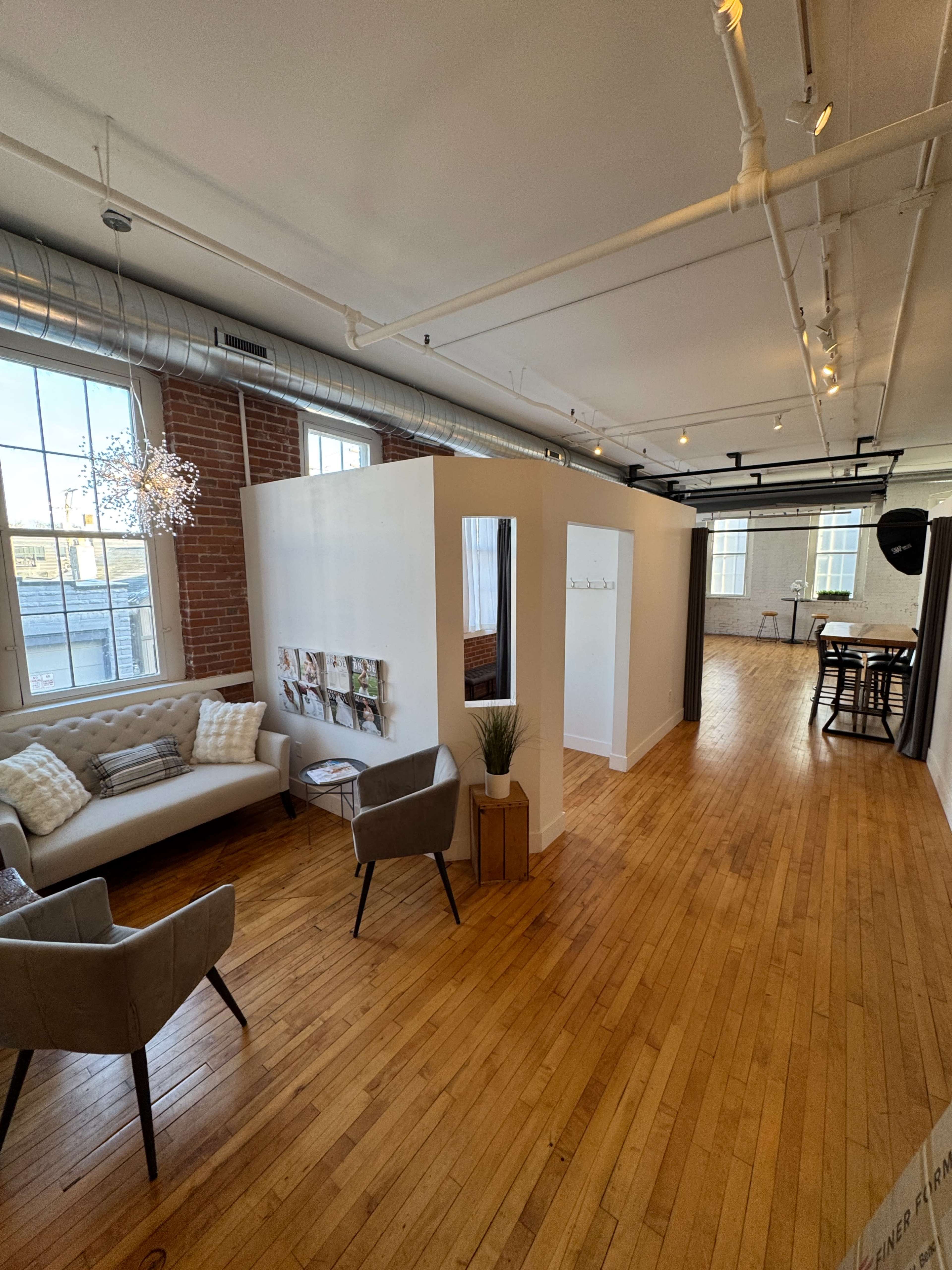 The image shows a bright, open office space featuring a seating area with a sofa and armchairs, a partition with a mirror, and a kitchenette in the background.