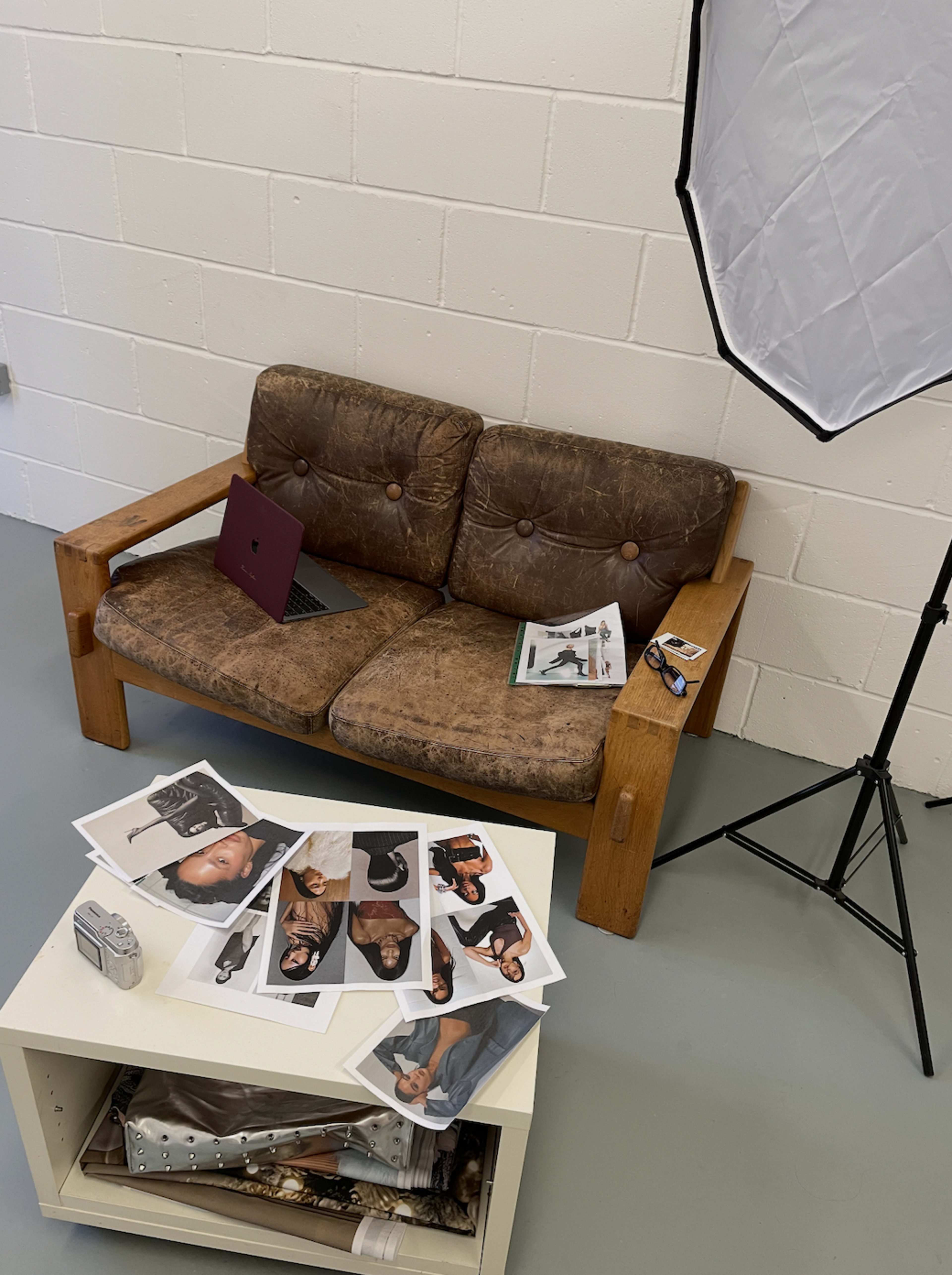A worn brown couch sits next to a white surface displaying printed photographs, with a laptop and magazines resting on the couch.