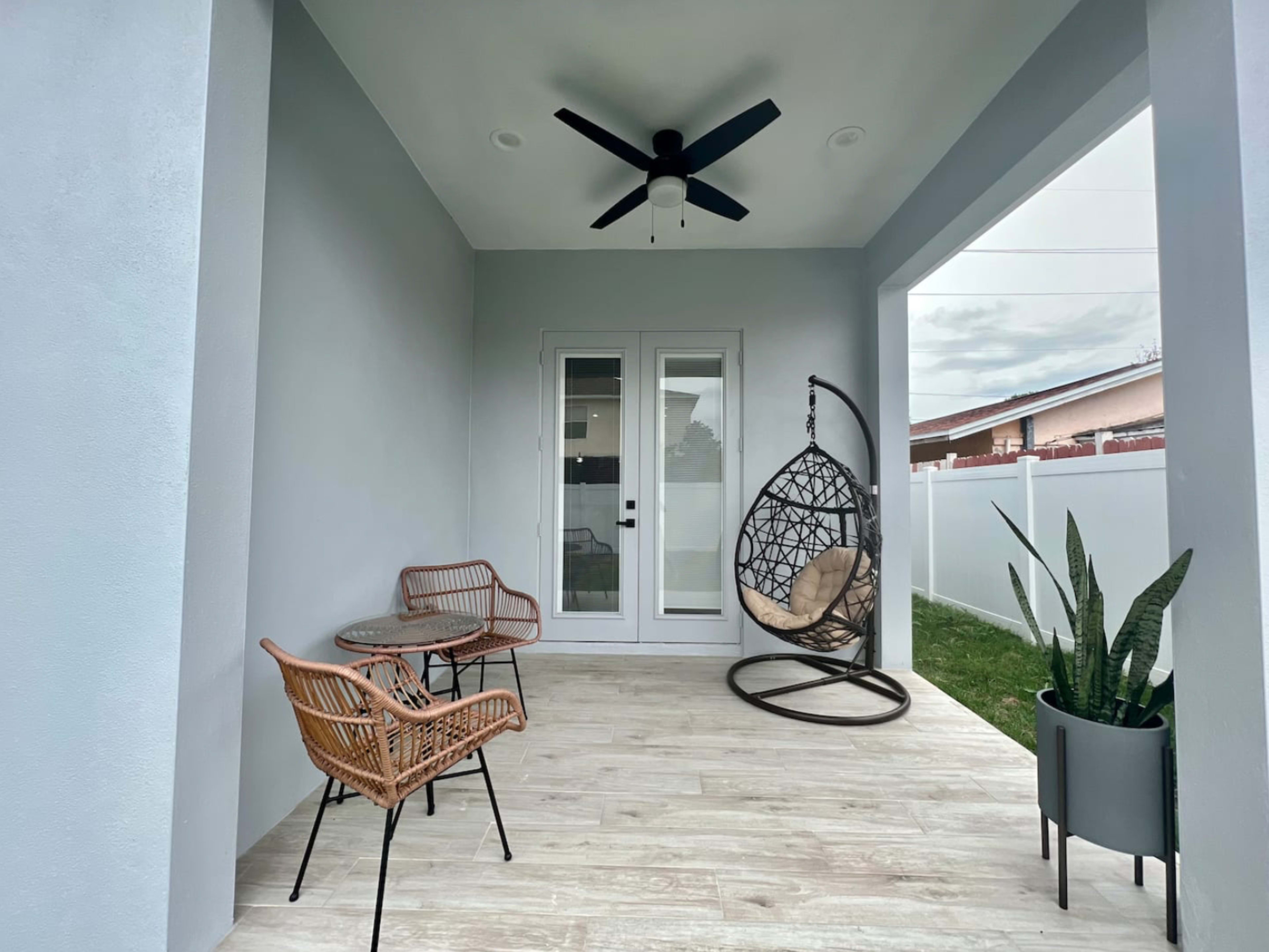 A covered outdoor patio features a hanging chair, two wicker chairs, a small table, and a potted plant.