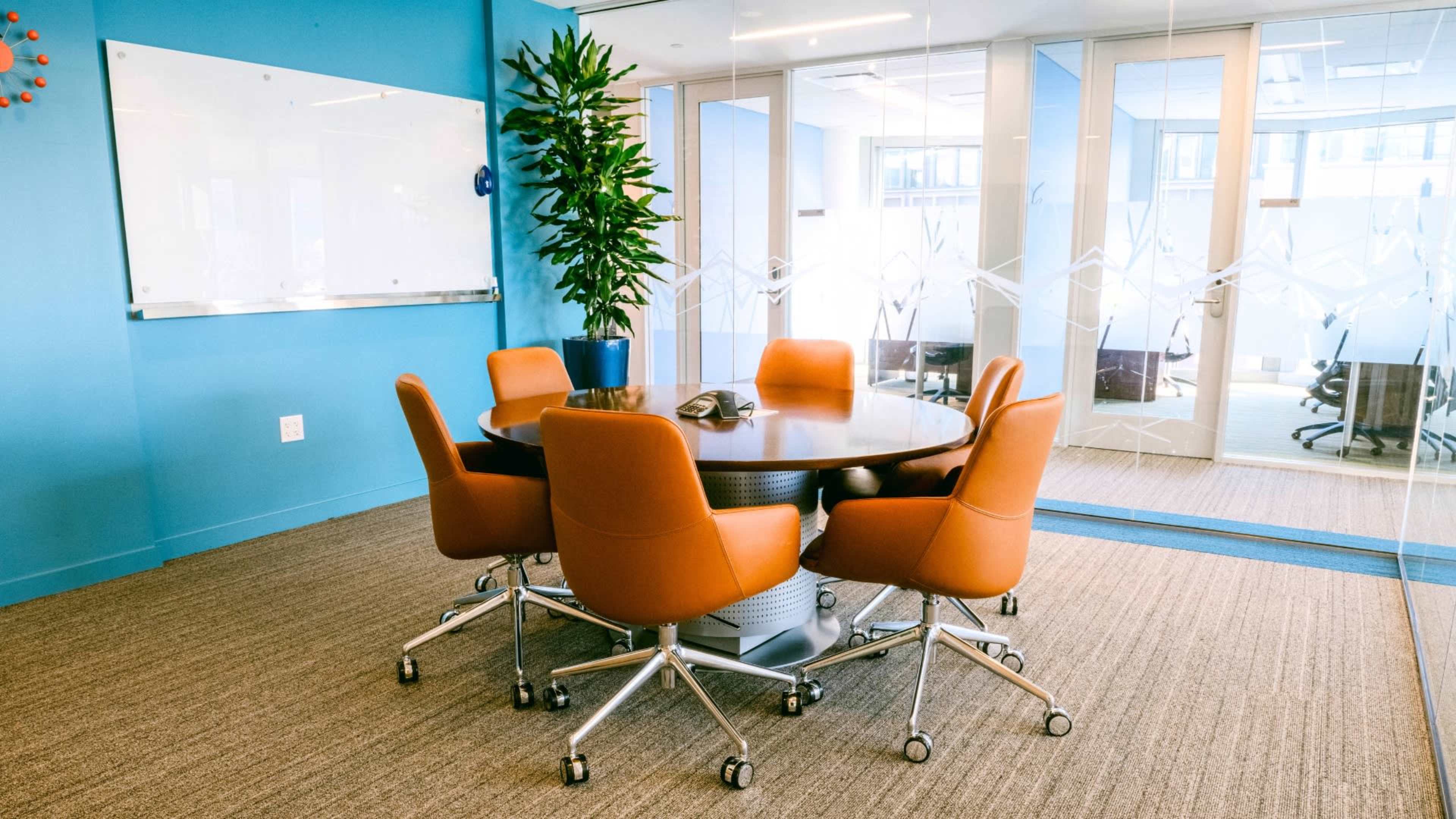 The image shows a conference room with a round table surrounded by four brown rolling chairs, a phone on the table, and a large plant in the corner.