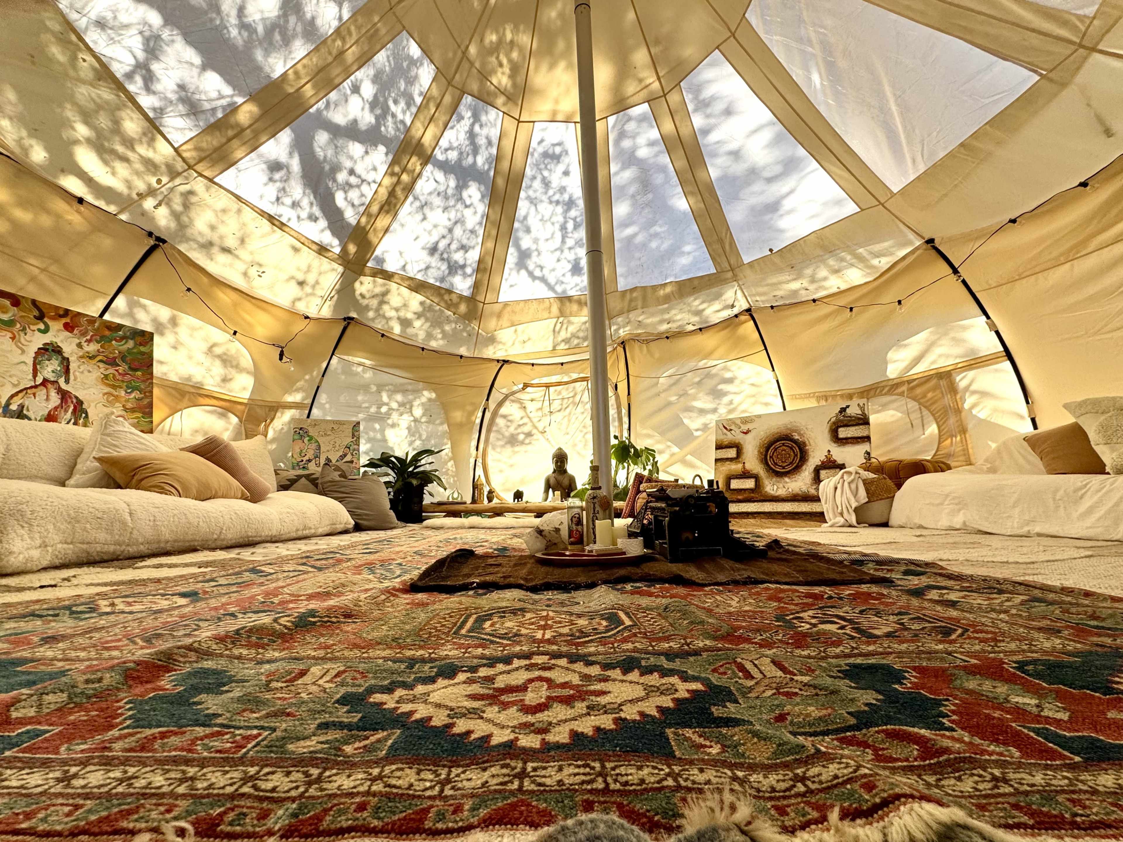 The interior of a spacious tent features a central area with a patterned rug, placed cushions, and art on the walls.