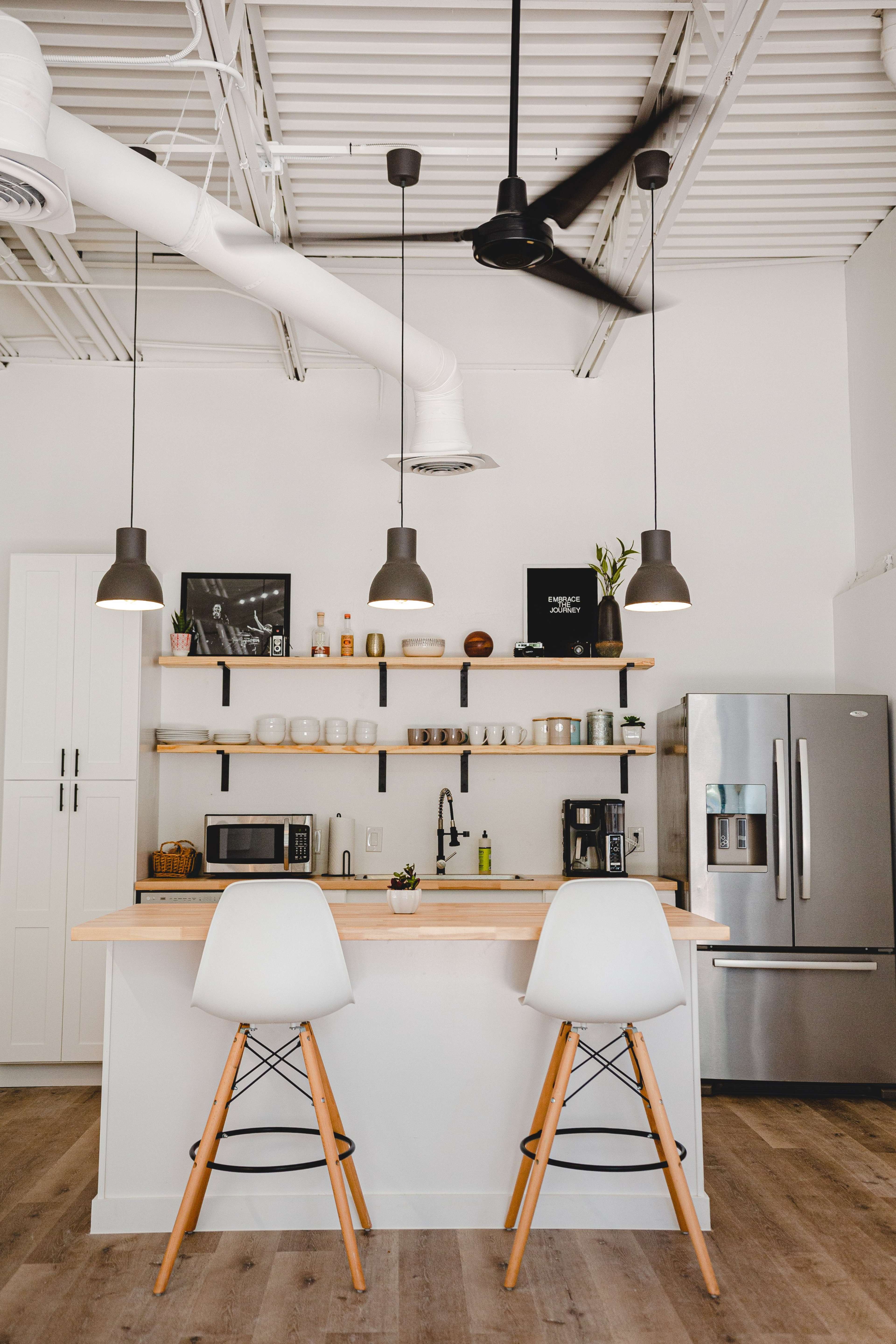A modern kitchen features a bar with two white stools, a wooden countertop, open shelves displaying various kitchen items, and stainless steel appliances.