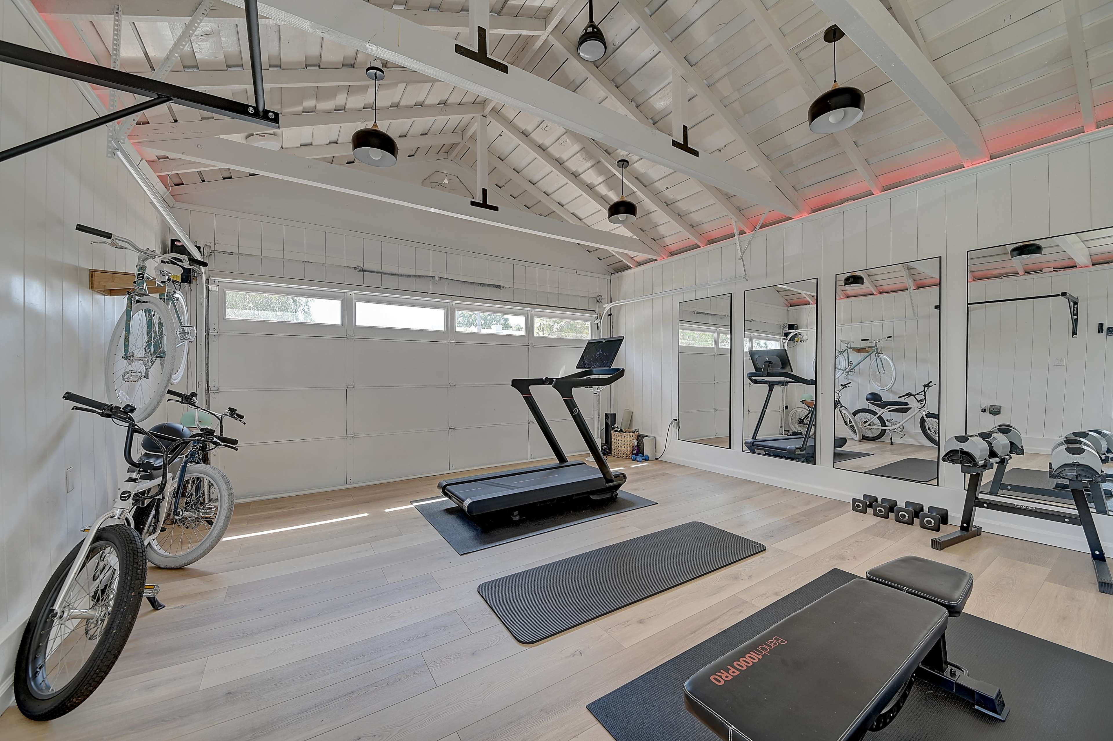 A modern home gym features a treadmill, free weights, and bicycles against a bright, airy backdrop with large mirrors and wooden beams.