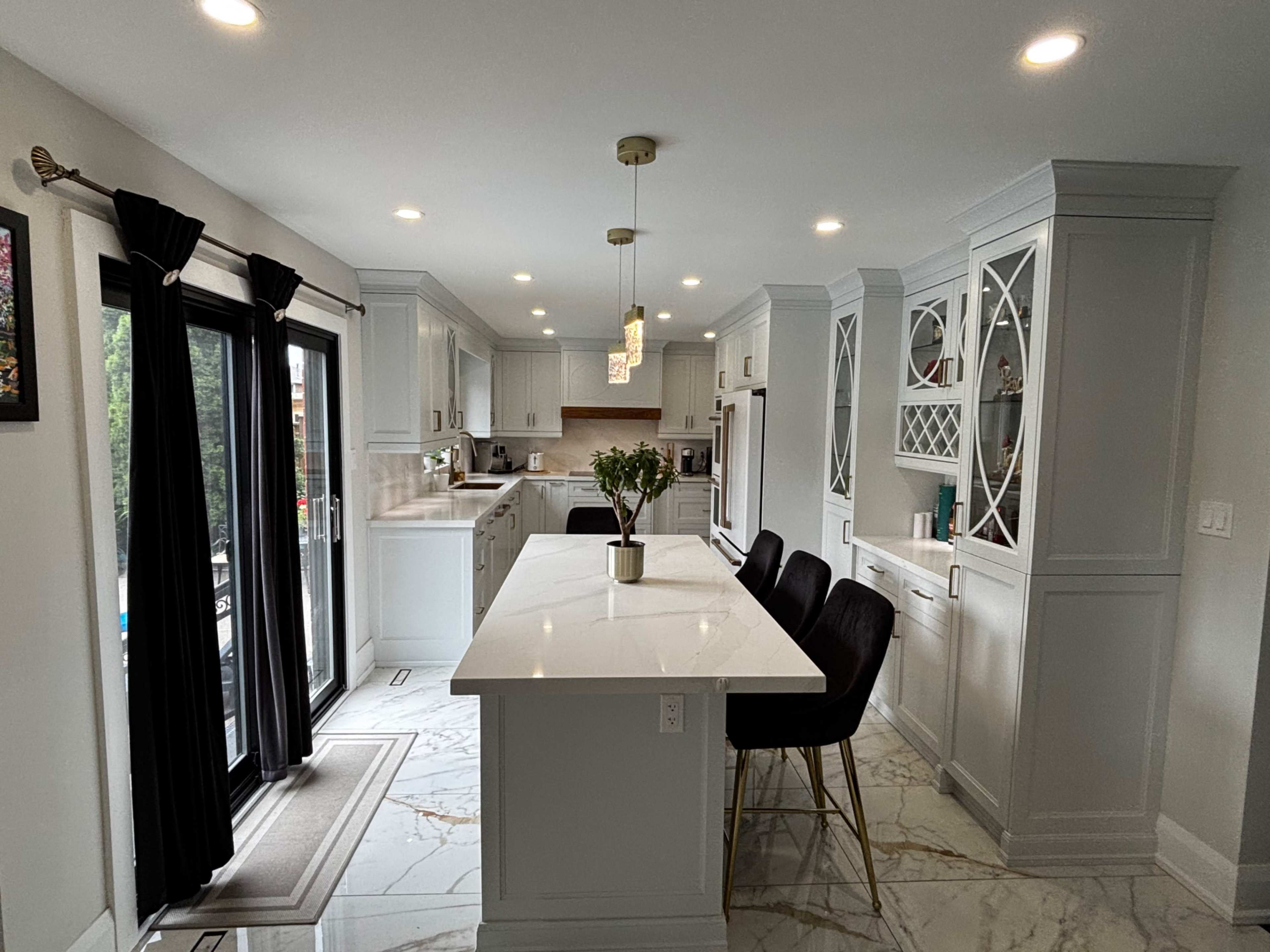 The image shows a modern kitchen with white cabinetry, a large island with bar stools, and large windows letting in natural light.
