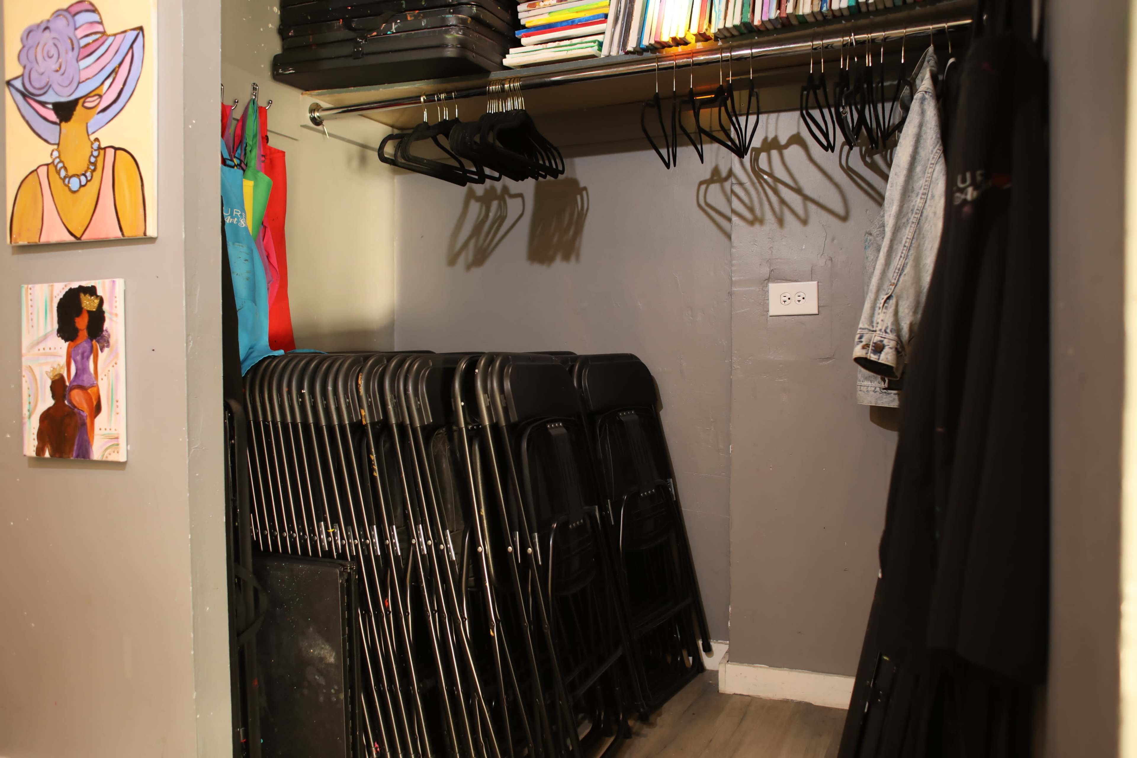 A closet with a stack of folding chairs, hangers on a rod, and artwork on the walls.