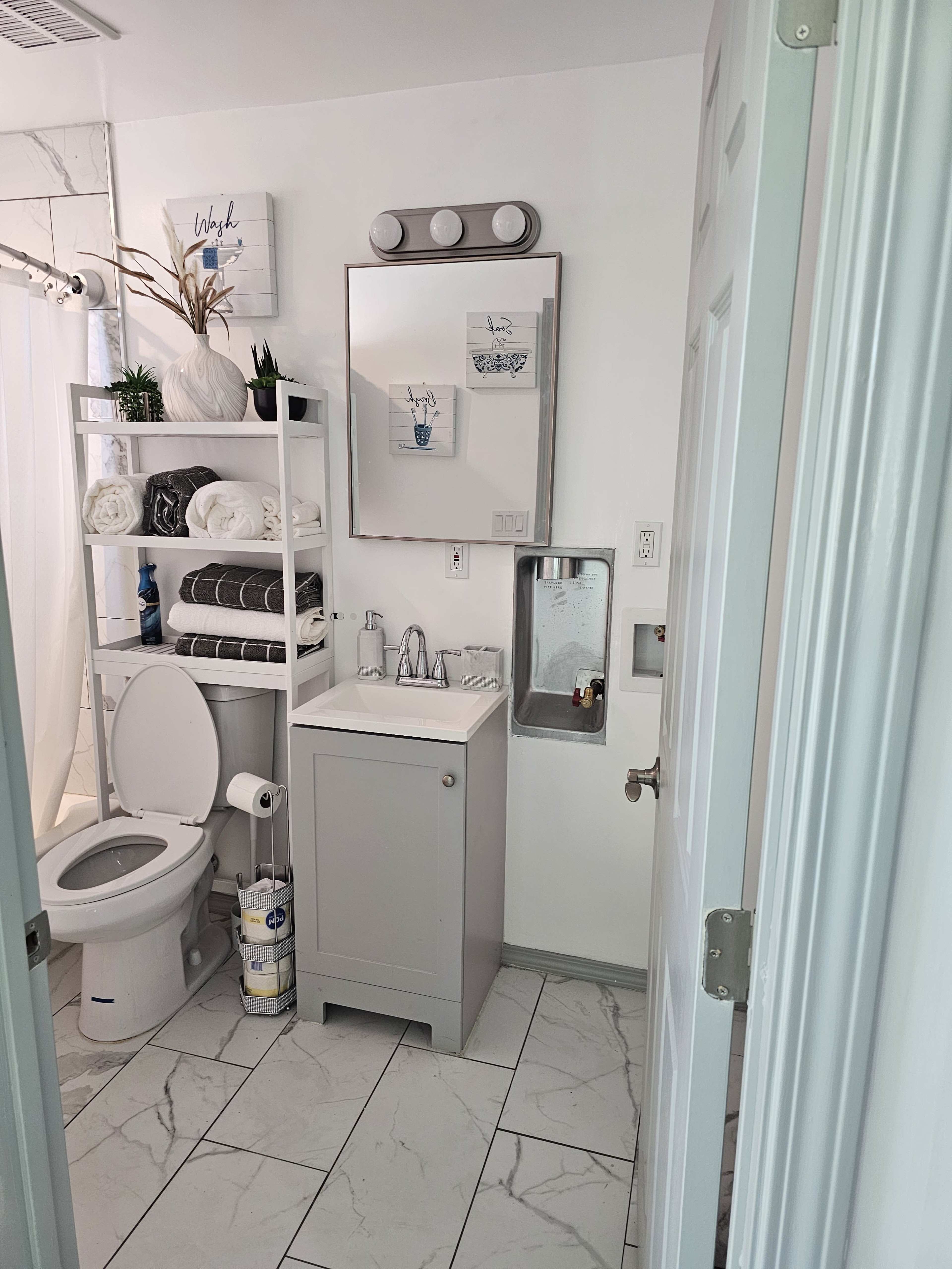 The image shows a clean bathroom with a toilet, a small sink with a mirror above it, a shelf with neatly arranged towels, and a large window allowing natural light.