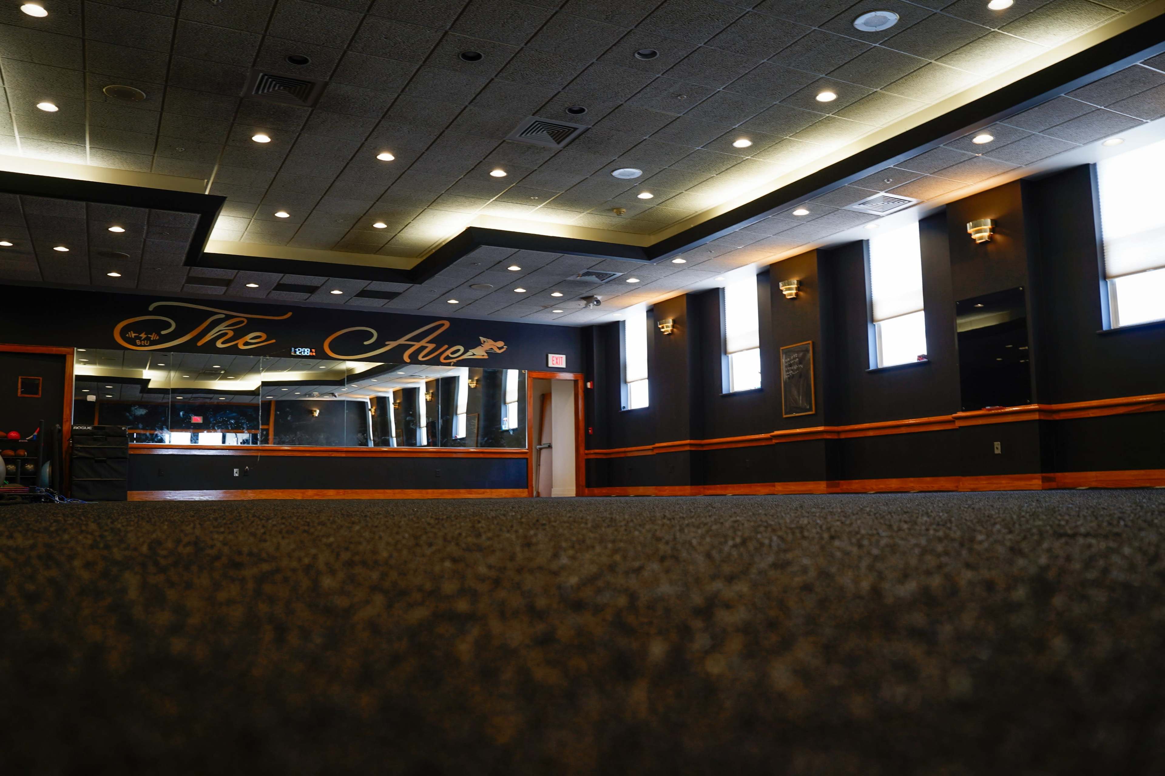 A spacious, empty room with carpeted flooring, featuring a mirror on one wall and the words "The Ave" painted above a wooden trim.