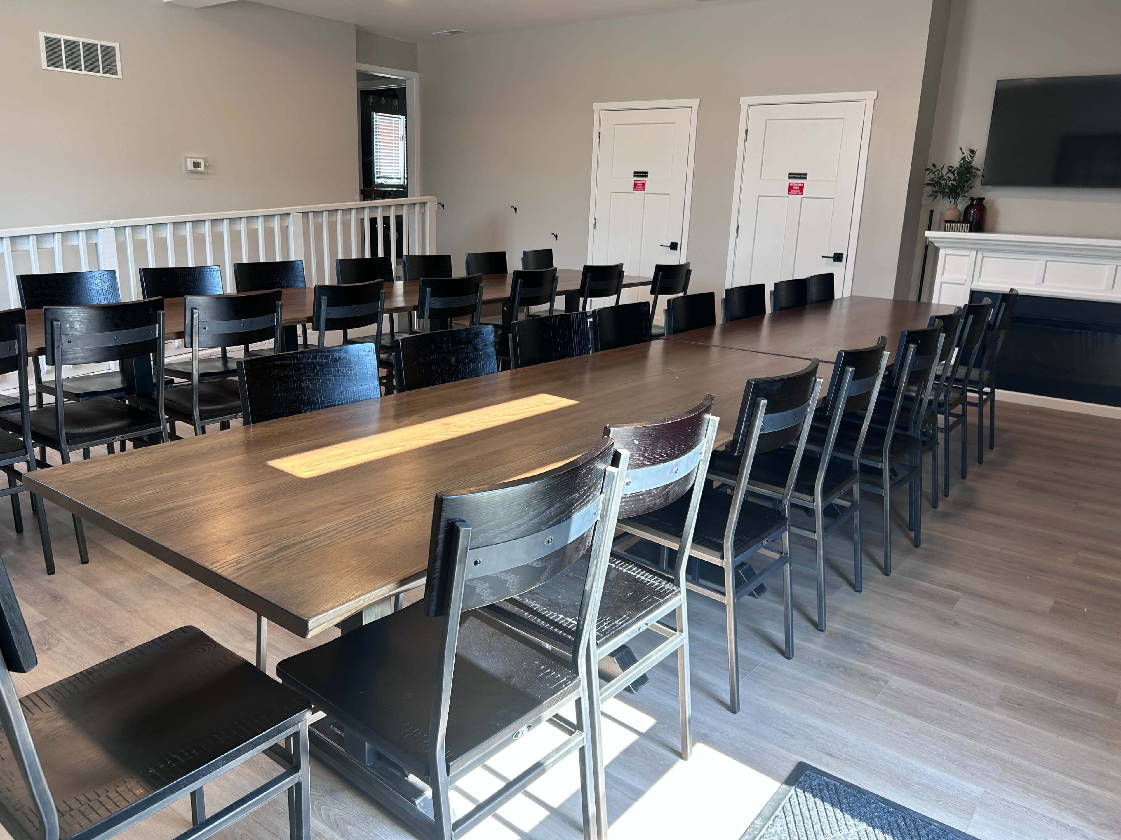 The image shows a large, rectangular wooden dining table surrounded by black chairs in a well-lit room with two closed doors and a television on the wall.