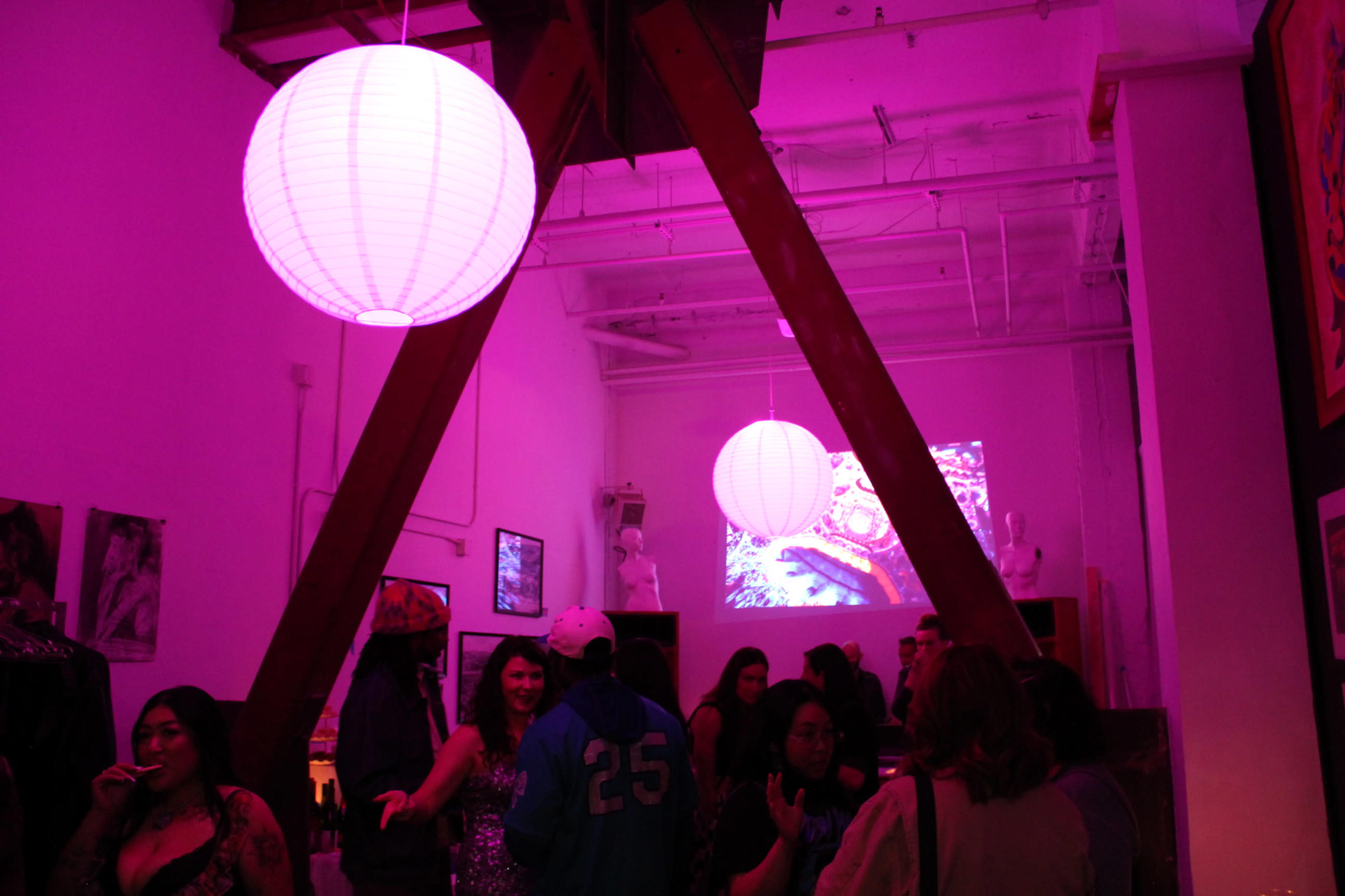 A group of people socialize under pink paper lanterns in a brightly lit room featuring wooden beams and a video projection on the wall.