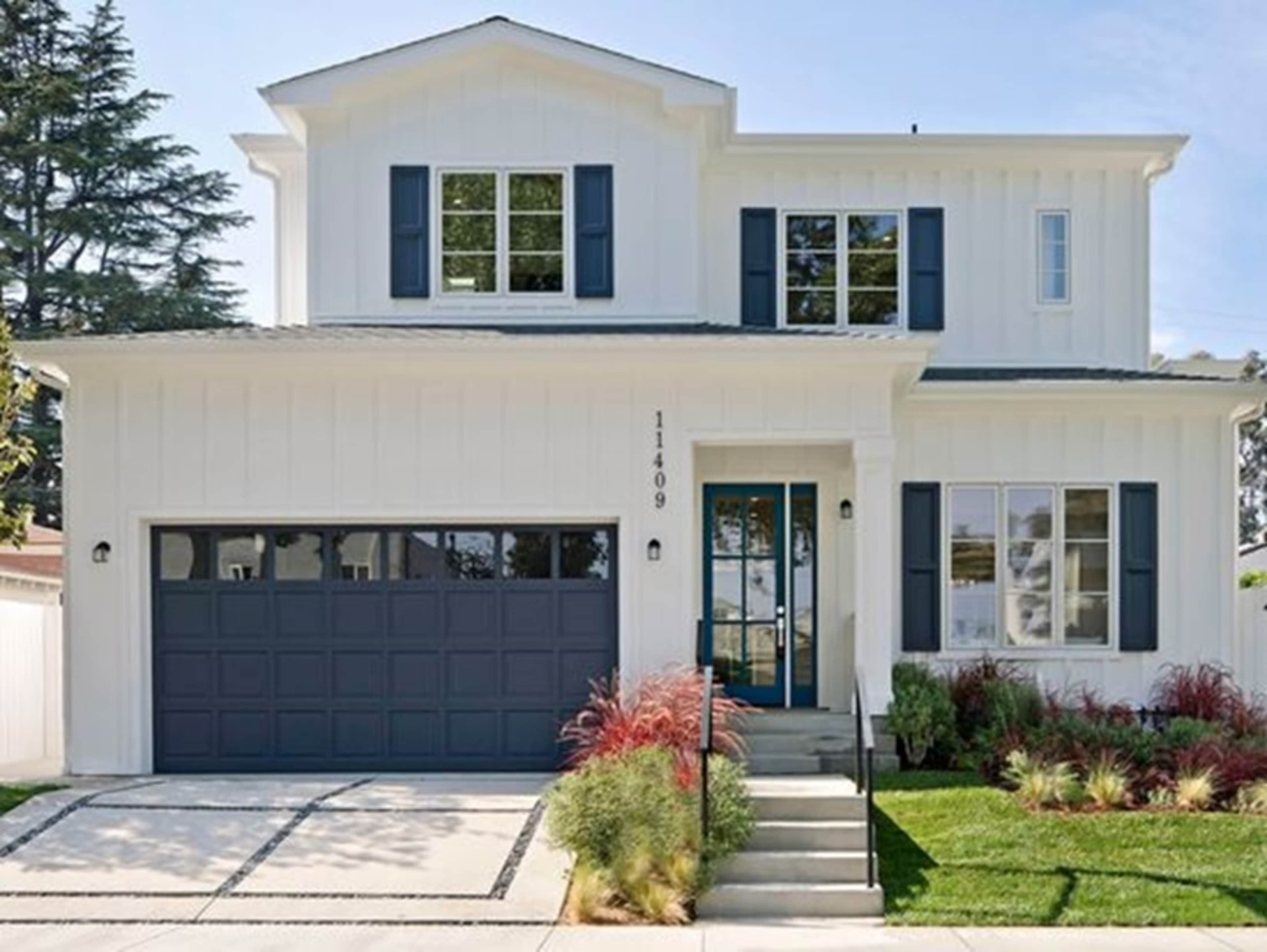 The image shows a two-story white house with a dark garage door and blue shutters, surrounded by neatly maintained landscaping.