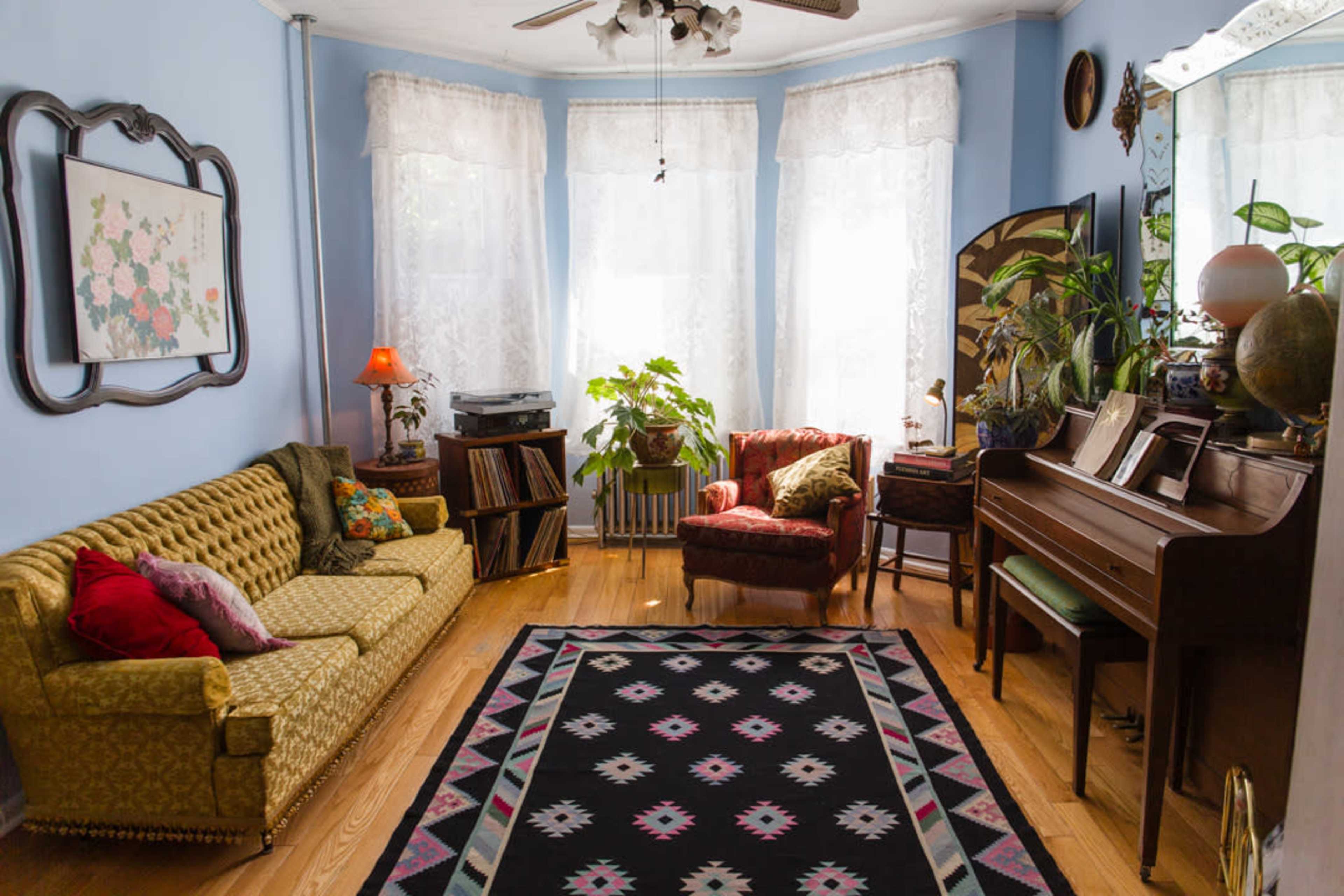 The image shows a cozy living room with large windows dressed in lace curtains, a green upholstered sofa, a red armchair, a piano, and various indoor plants.