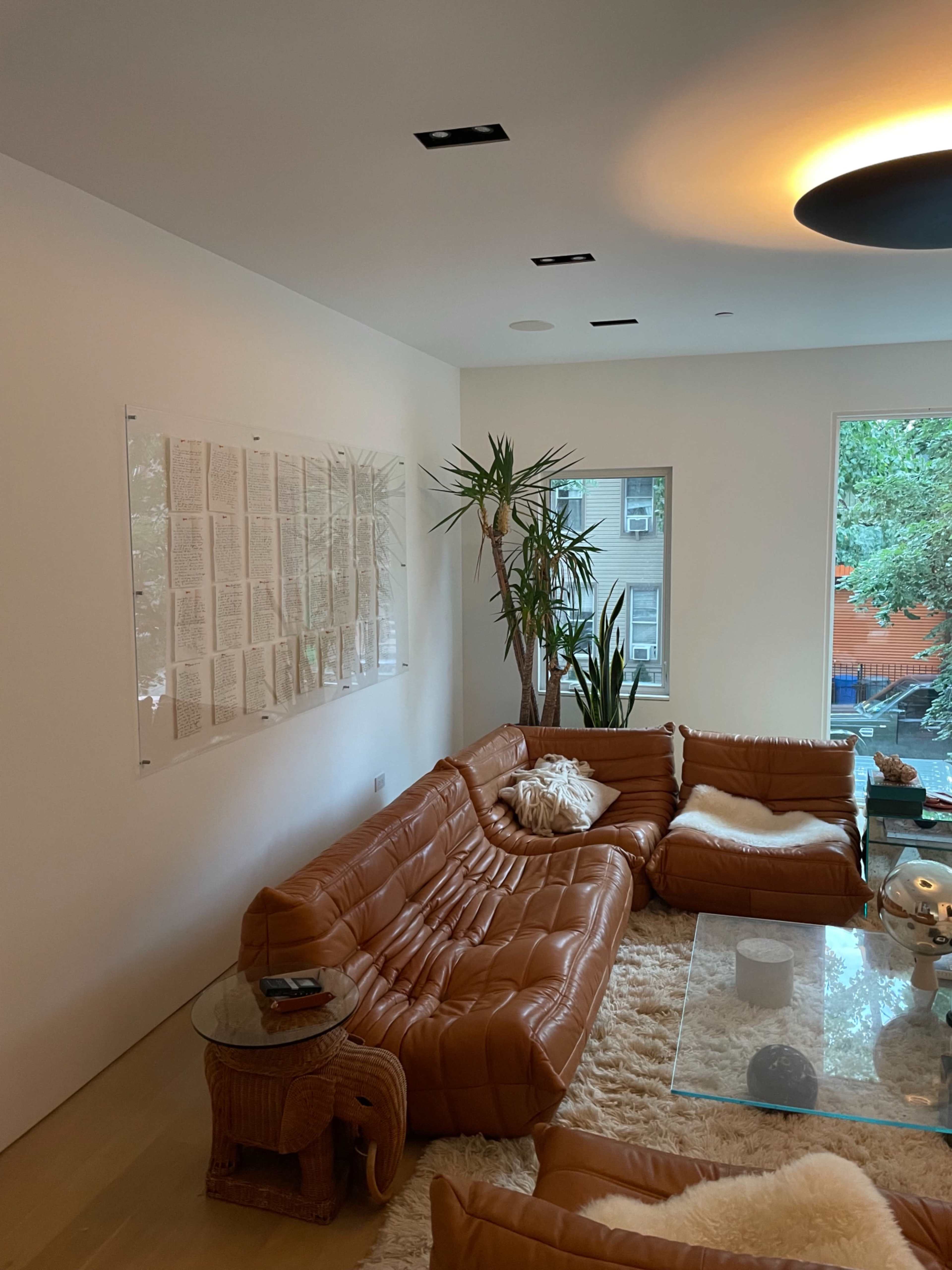 A modern living room features a brown leather sectional sofa, a glass coffee table, and a wall adorned with a grid of framed documents.