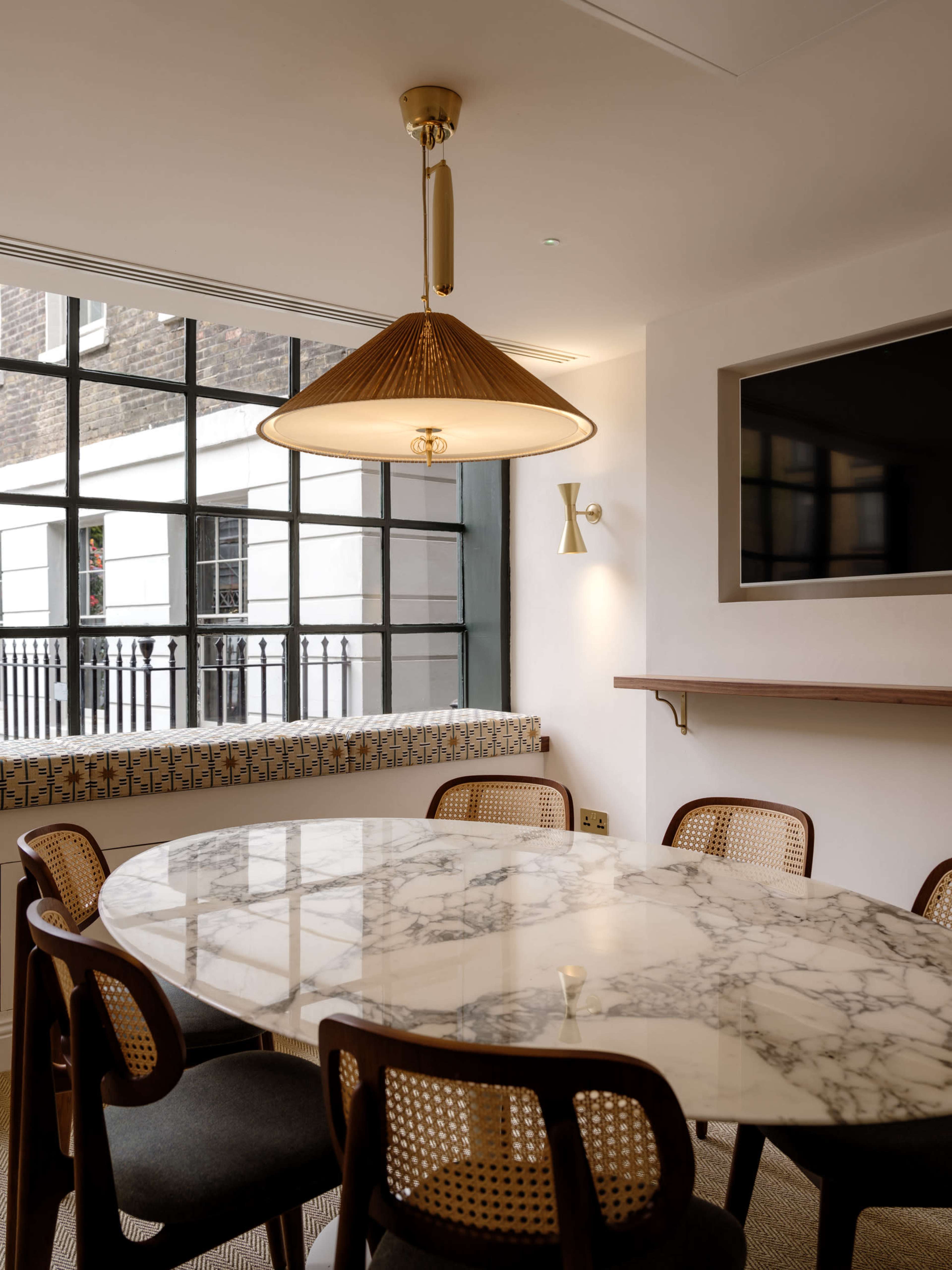 A modern dining area features a marble table surrounded by wooden chairs and illuminated by a pendant light, with large windows providing an outdoor view.