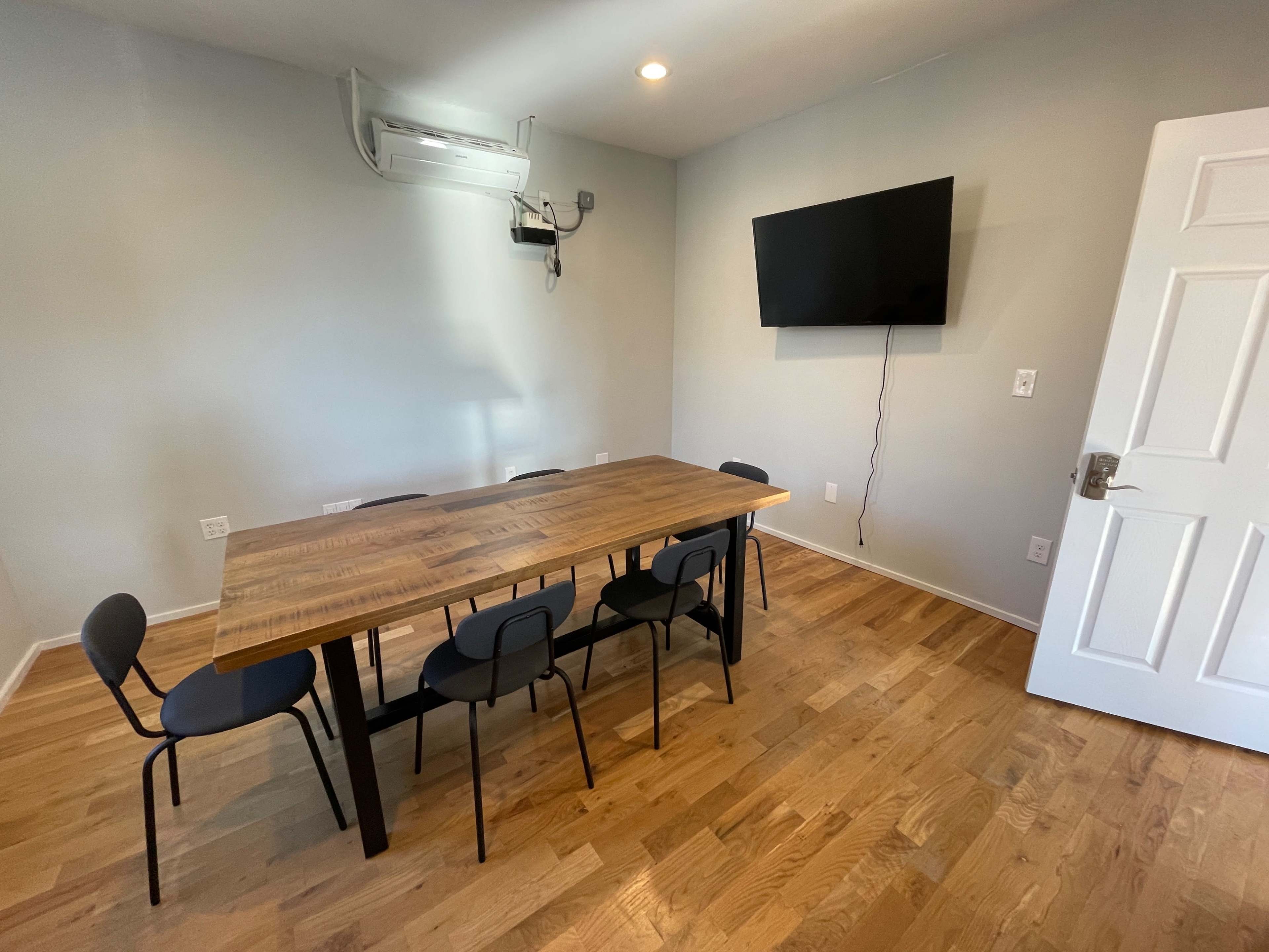 A modern dining area with a wooden table and six black chairs, a wall-mounted television, and light-colored walls and flooring.