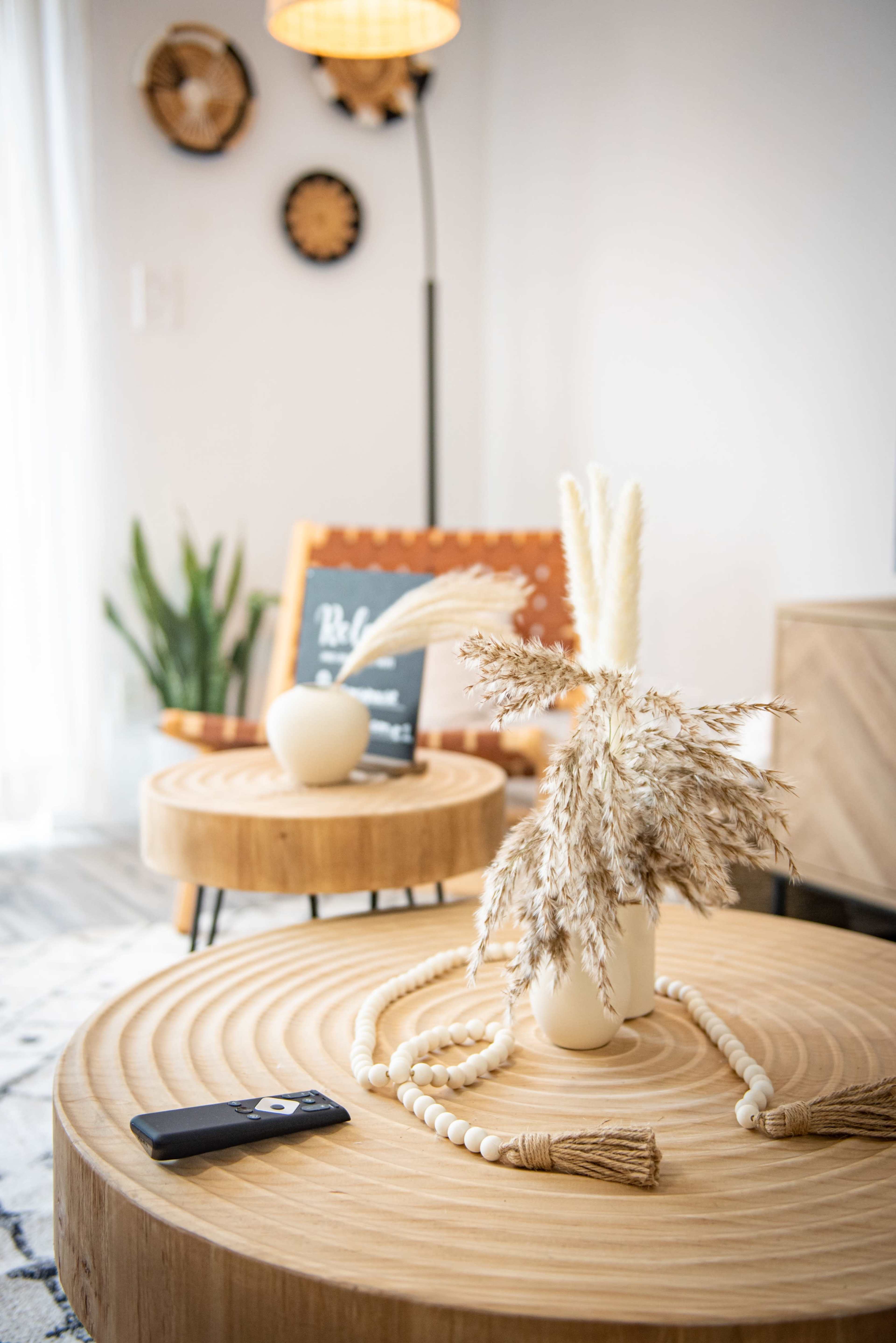 A wooden coffee table holds a vase of dried flowers and beads, with a remote control beside it, while a patterned chair and a plant are visible in the background.