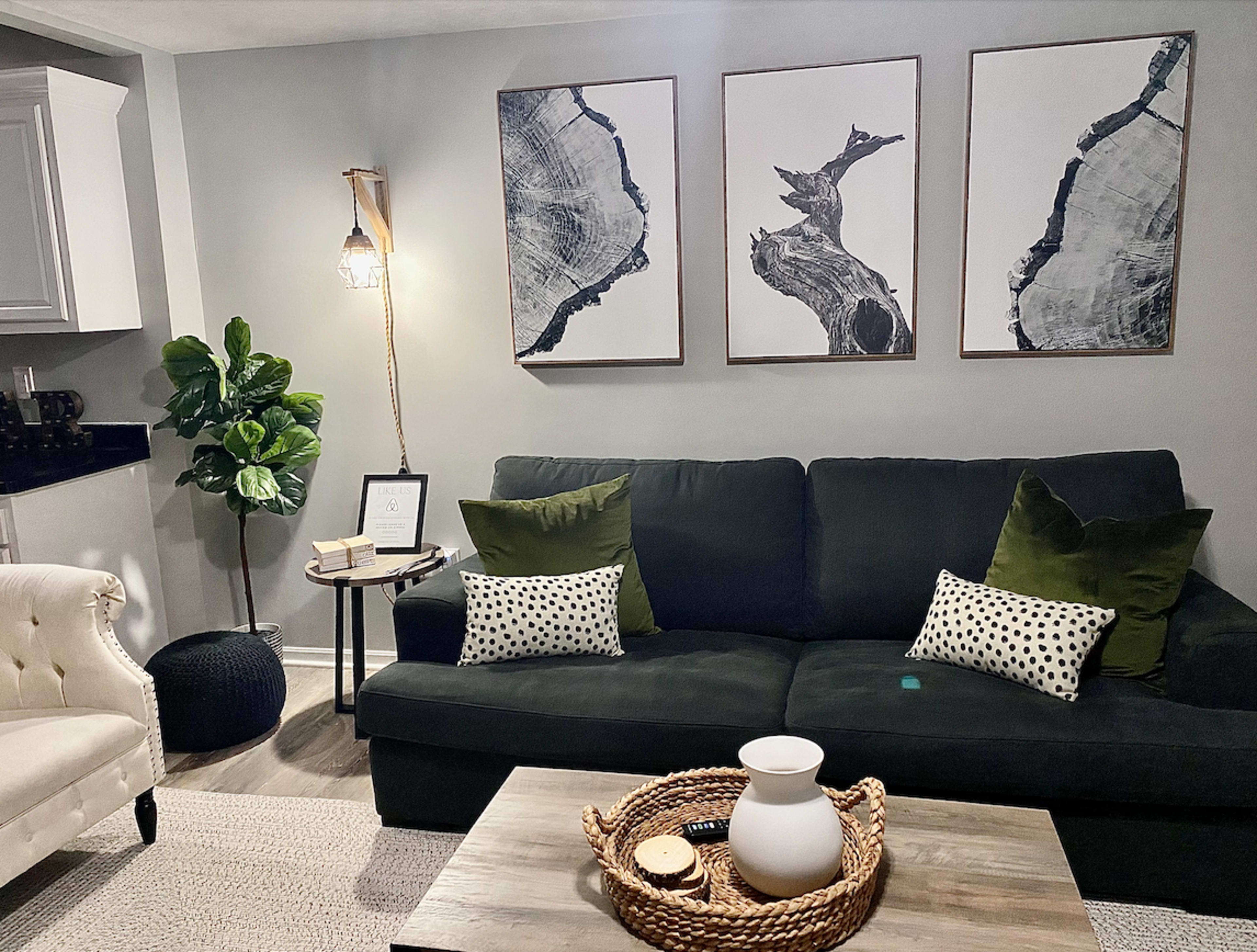 A modern living room featuring a dark sofa with decorative pillows, three framed black-and-white tree art pieces on the wall, a lamp, a potted plant, and a coffee table with a woven basket and a ceramic pitcher.