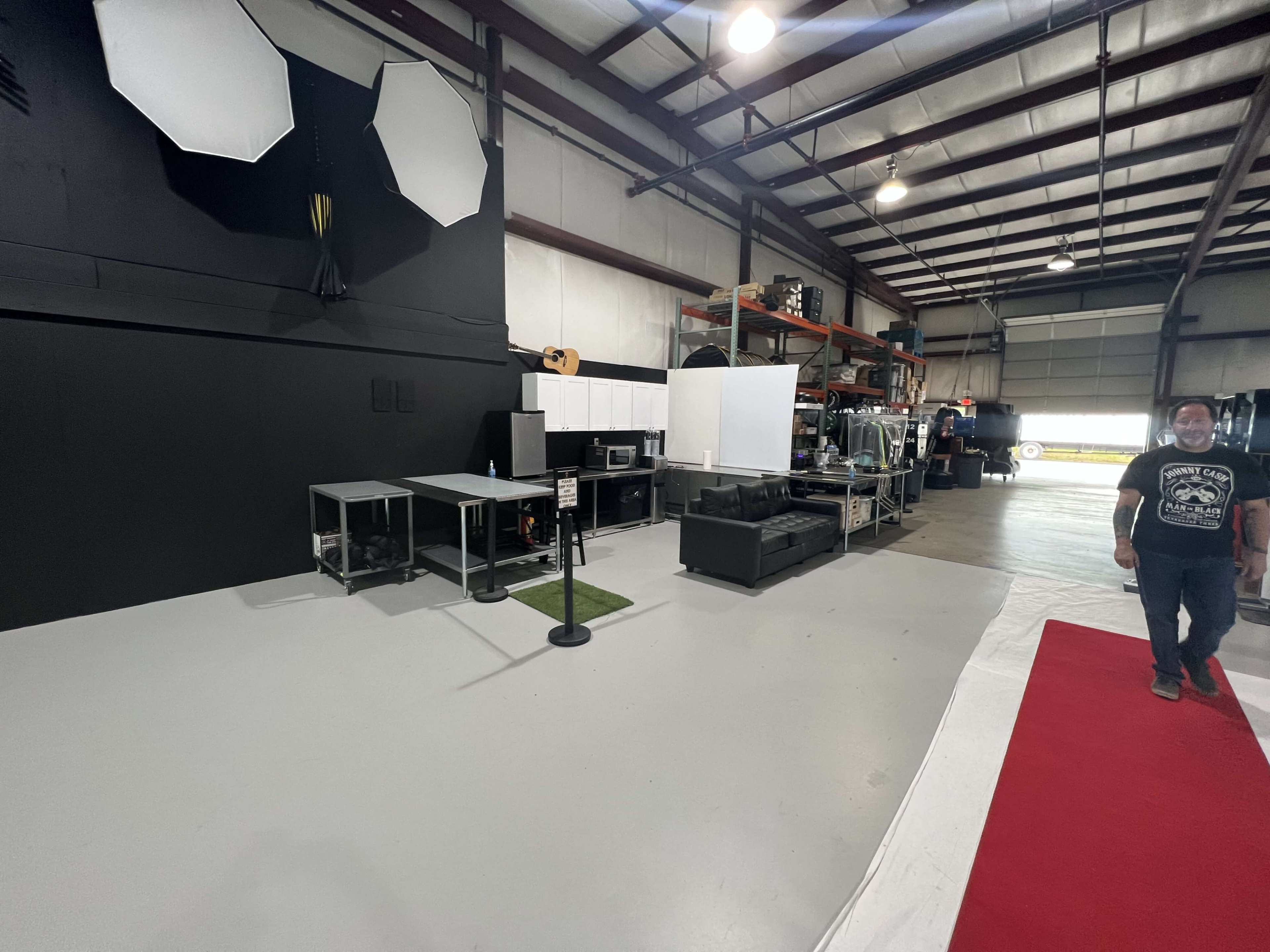 The image shows an industrial space with a grey floor, a black wall, several storage shelves filled with equipment, and a person standing near a red carpet.