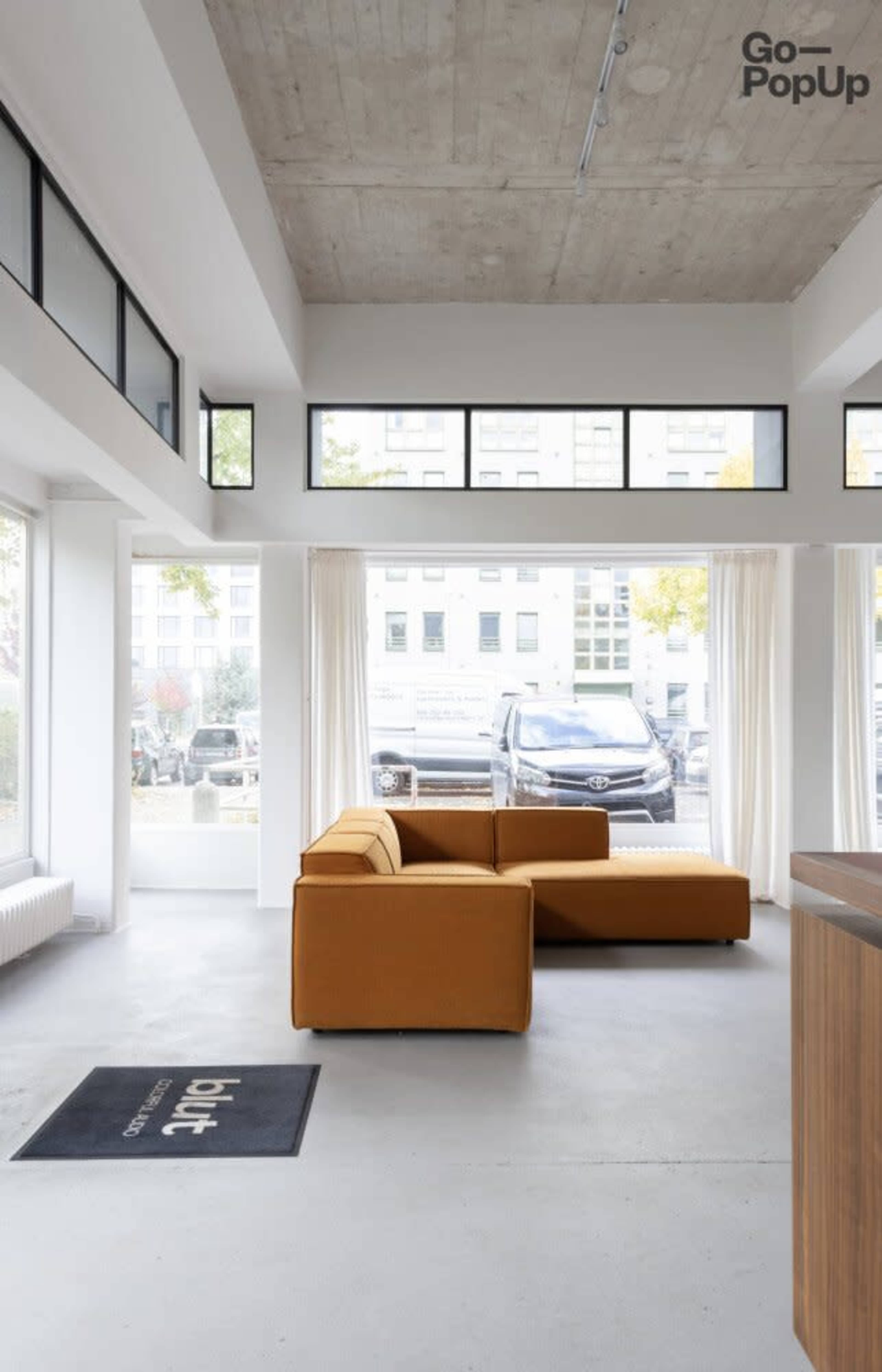 The image shows a modern, minimalistic interior with a brown couch facing large windows, allowing natural light to flood the space.