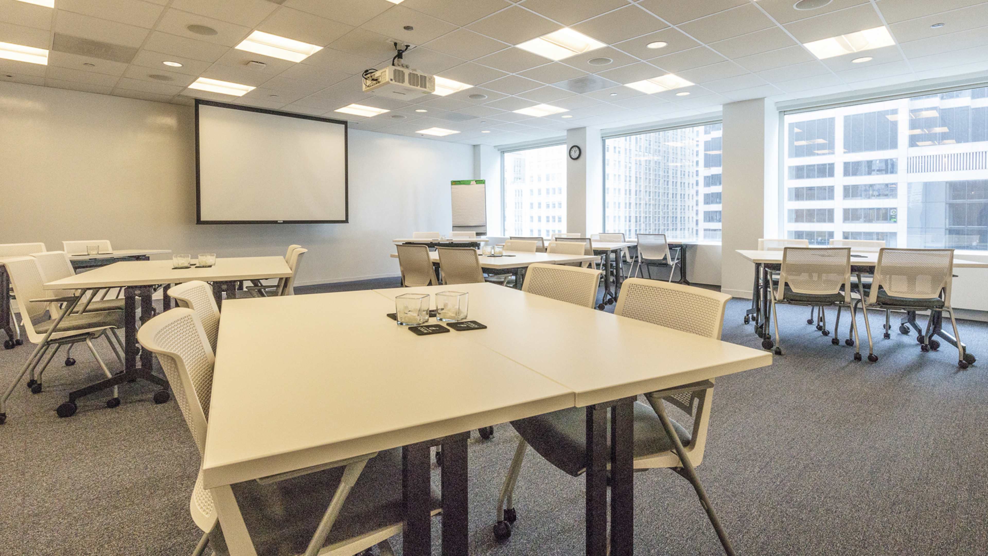 A modern conference room with several white tables and rolling chairs, a large screen at the front, and bright windows overlooking a cityscape.
