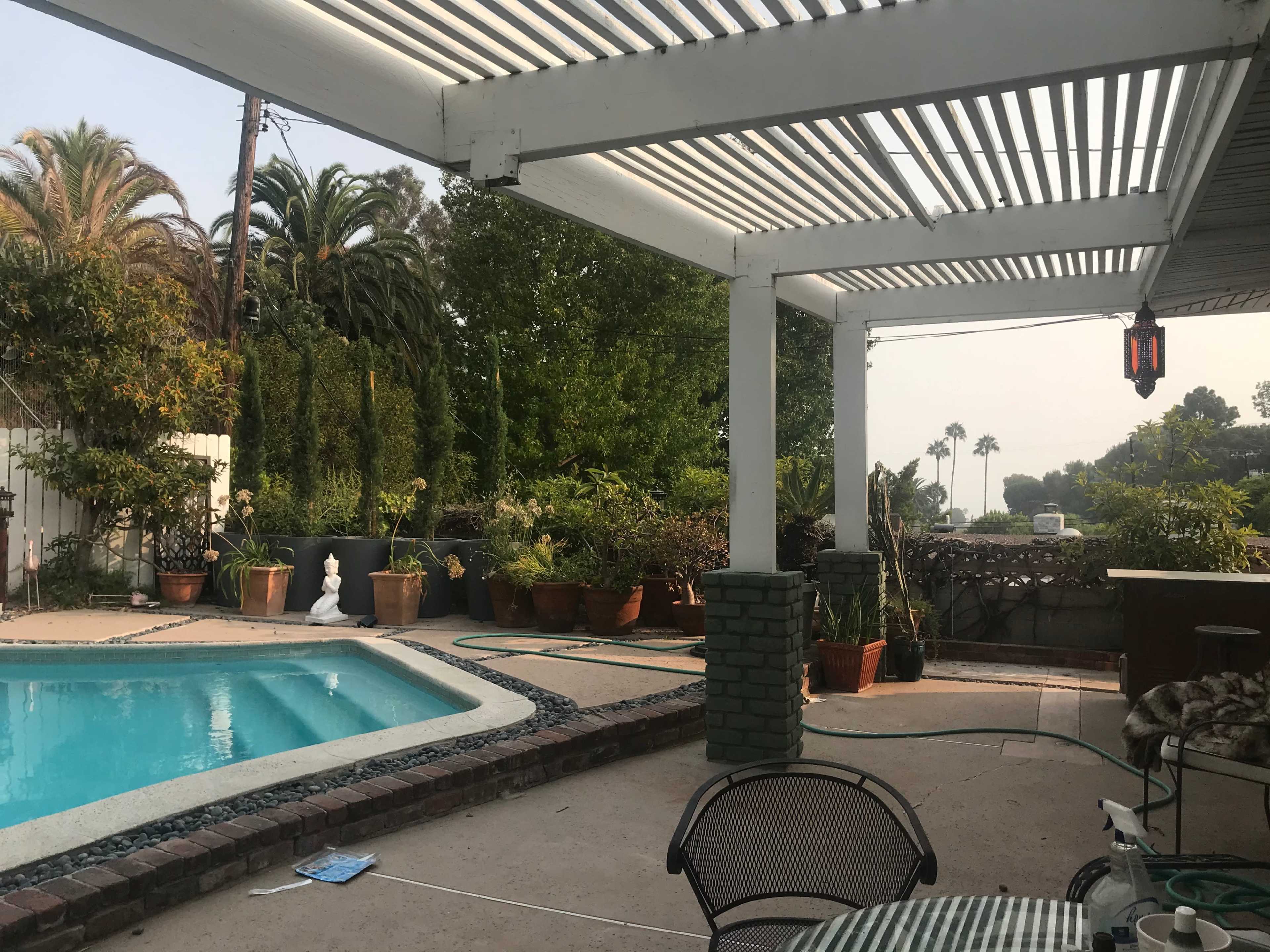 A patio with a swimming pool is surrounded by potted plants and palm trees under a partially shaded pergola.