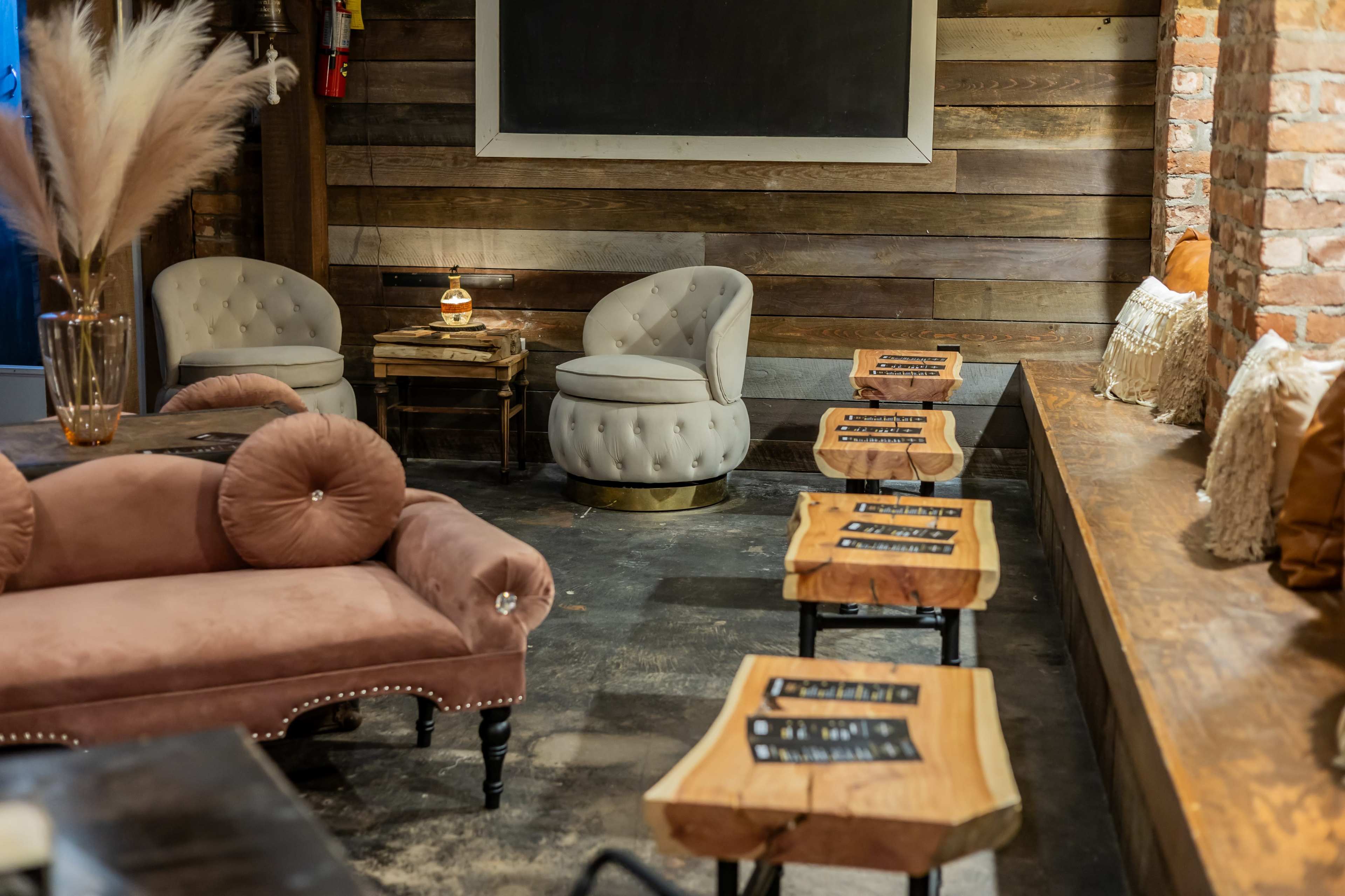 The image shows a cozy interior space with a tufted pink sofa, wooden stools, and a rustic brick wall, illuminated by soft lighting.