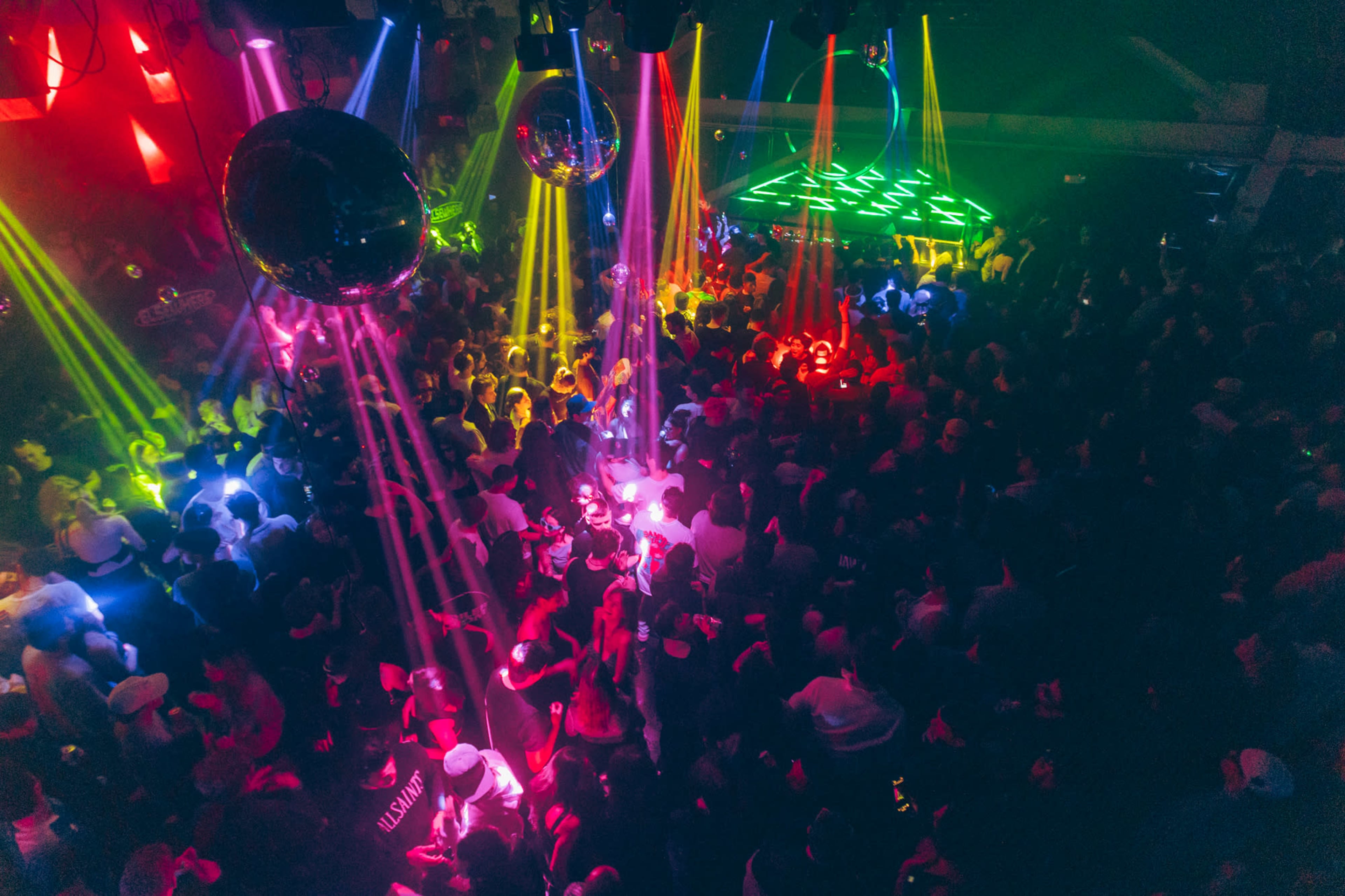 A crowded nightclub scene is illuminated by colorful lights and reflective disco balls.