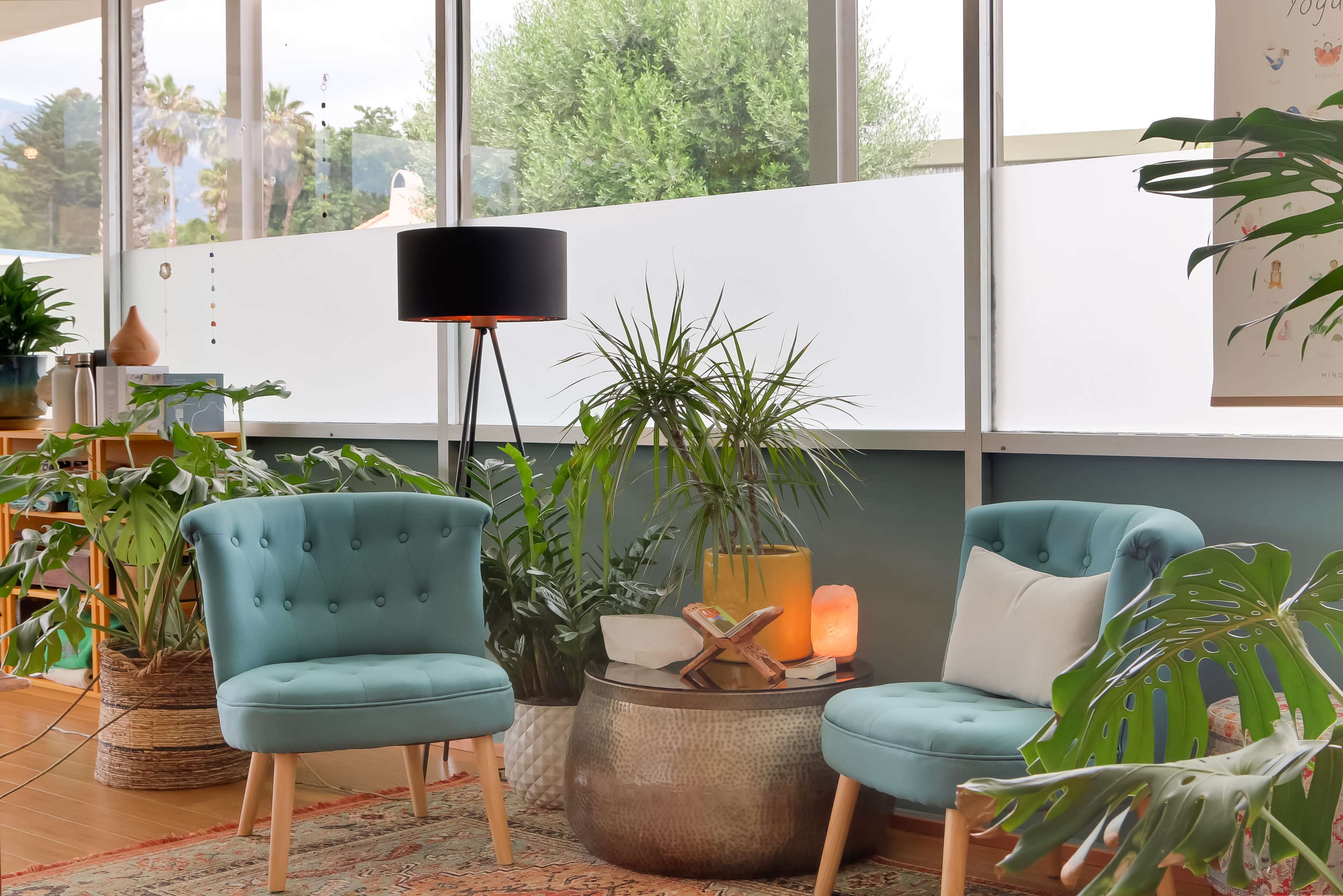 The scene features two light blue chairs flanking a round metallic table, surrounded by plants and illuminated by a floor lamp in a modern, light-filled room.