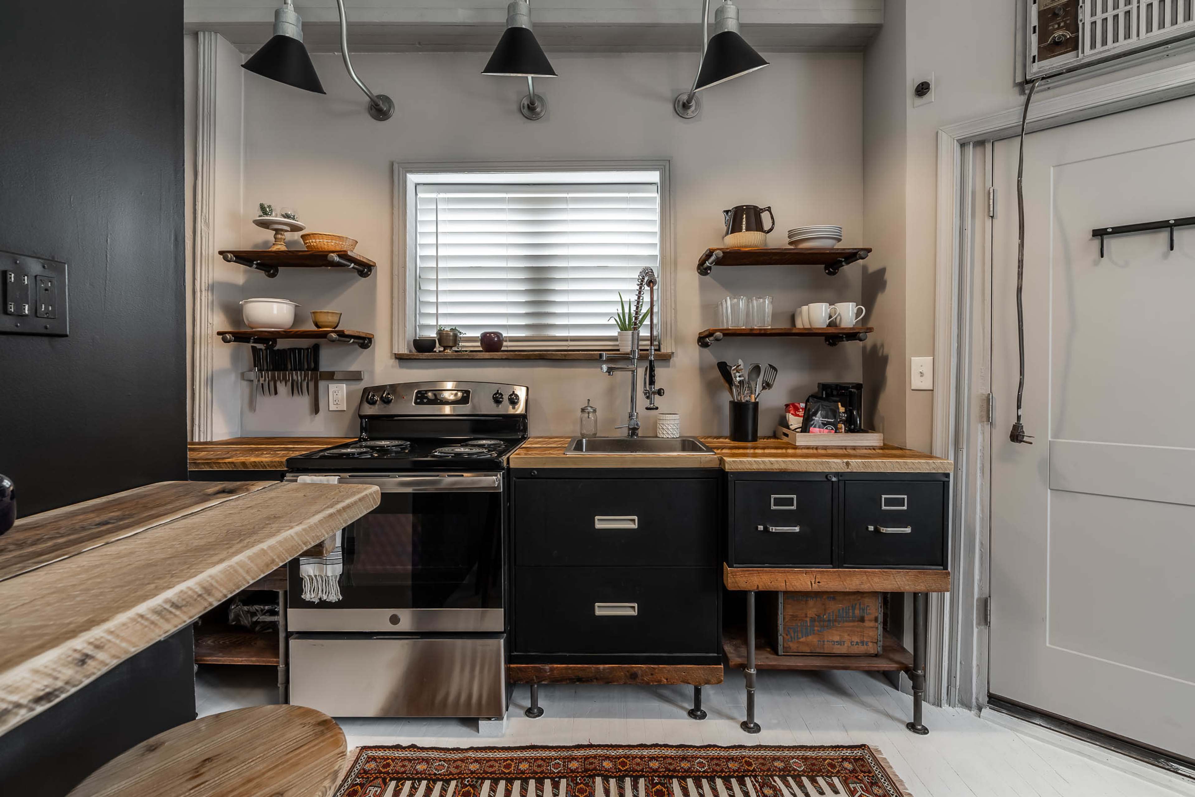A compact kitchen with black cabinets, wooden shelves, a stainless steel stove, and a farmhouse-style sink.