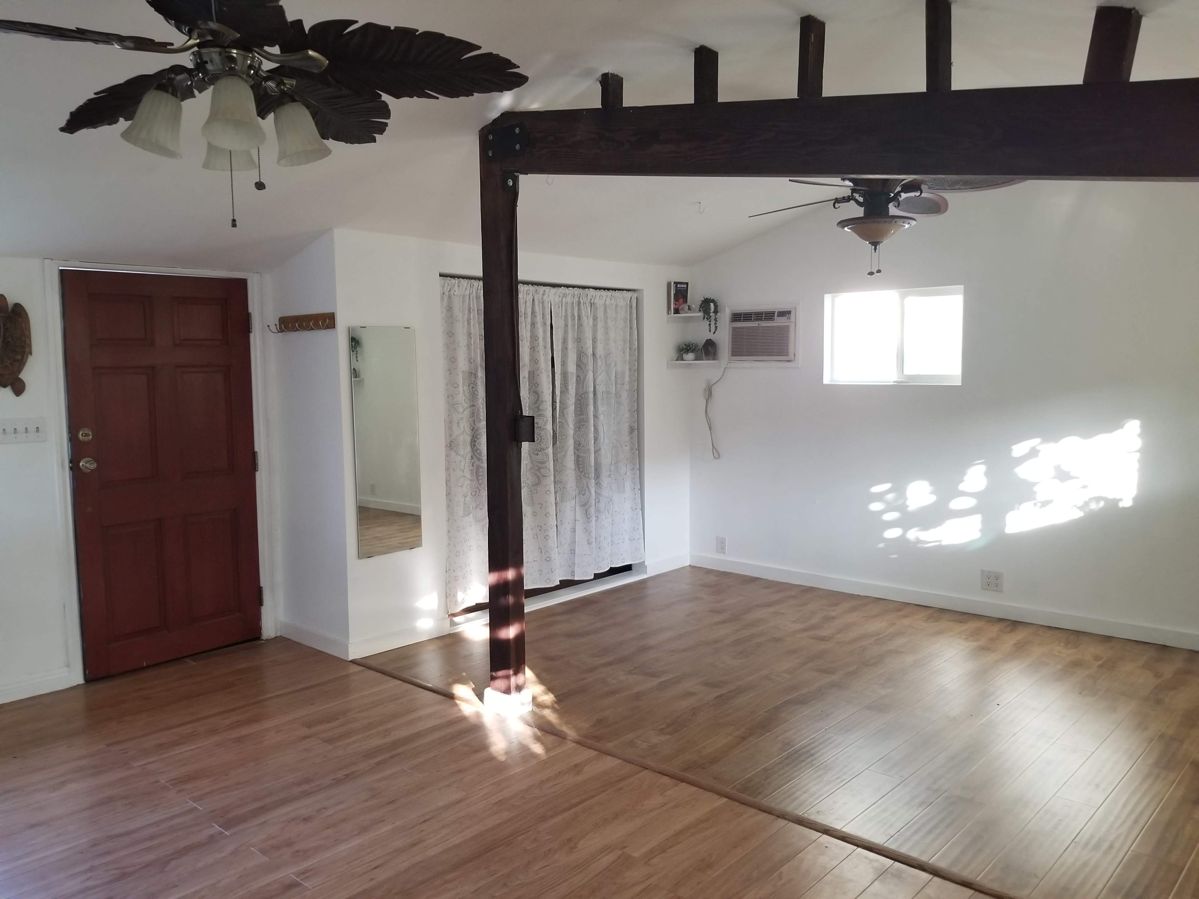 The image shows a room with wooden flooring, a mirror, a red front door, and a ceiling fan with palm leaf blades.