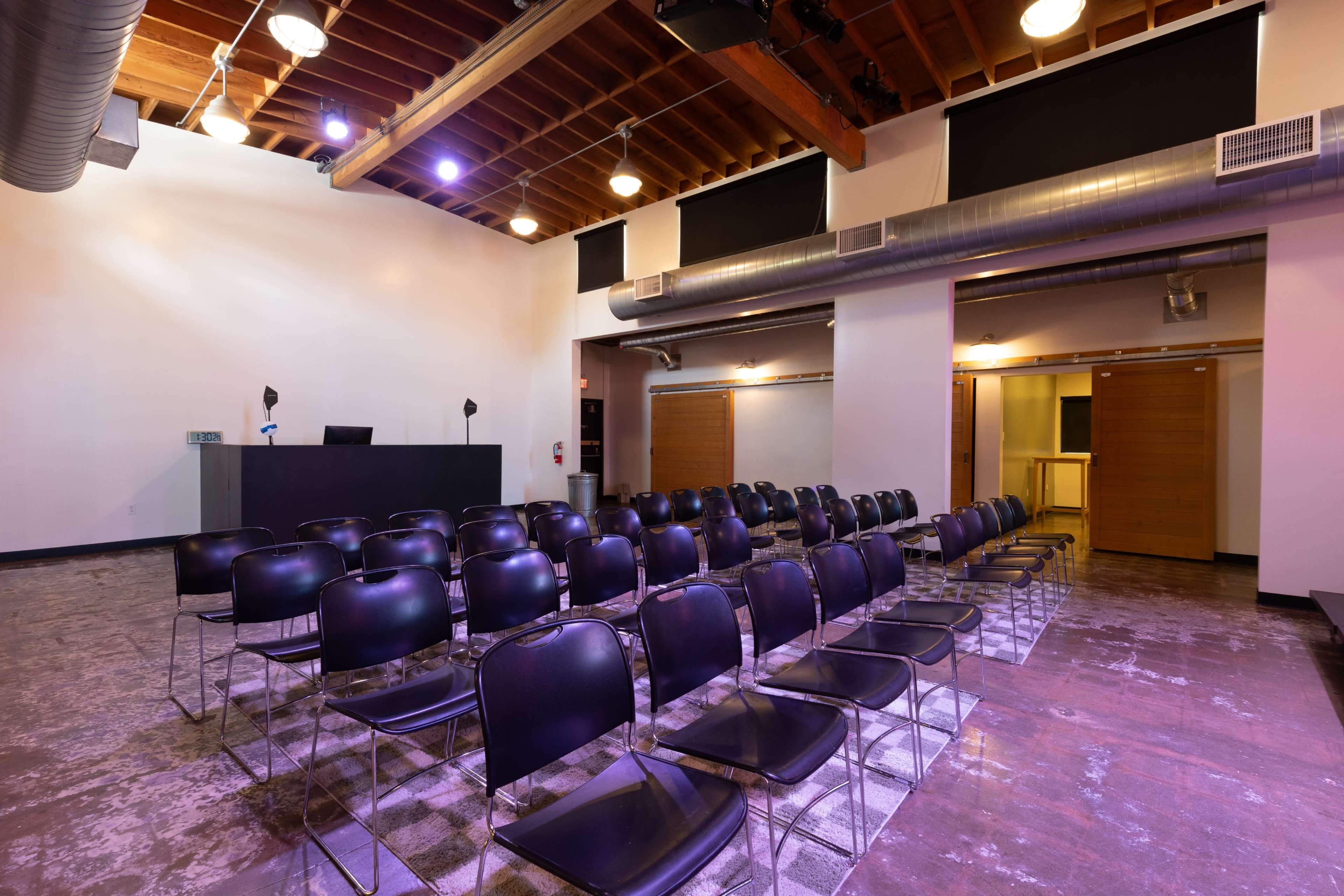 The image shows an empty auditorium with rows of black chairs facing a low stage and a backdrop of exposed wooden beams and industrial-style lighting.