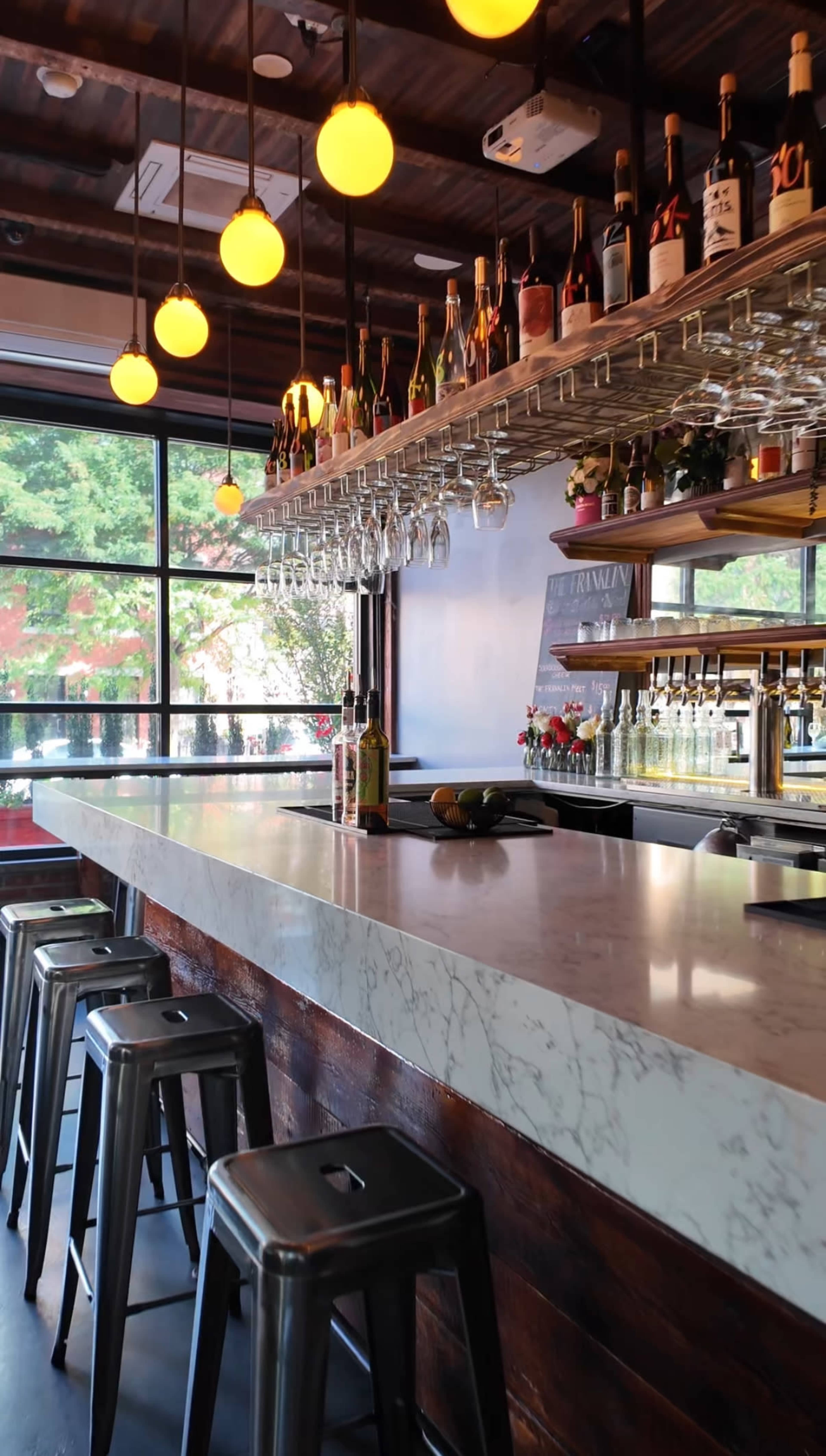 The image shows a modern bar with a marble countertop, metal stools, and shelves stocked with bottles and glasses, set against large windows overlooking greenery.