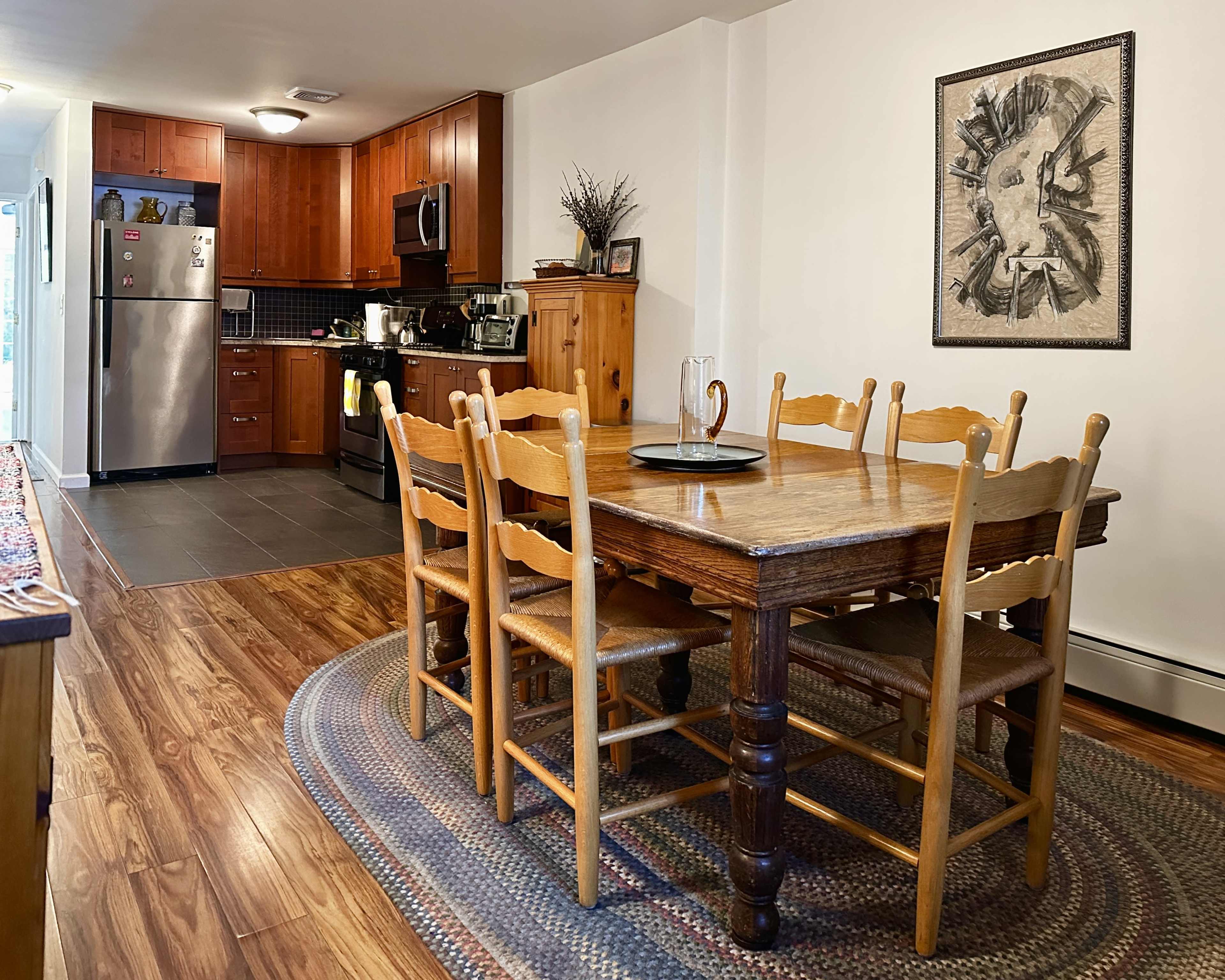 The image shows a dining area featuring a wooden table surrounded by six chairs, with an open kitchen visible in the background.
