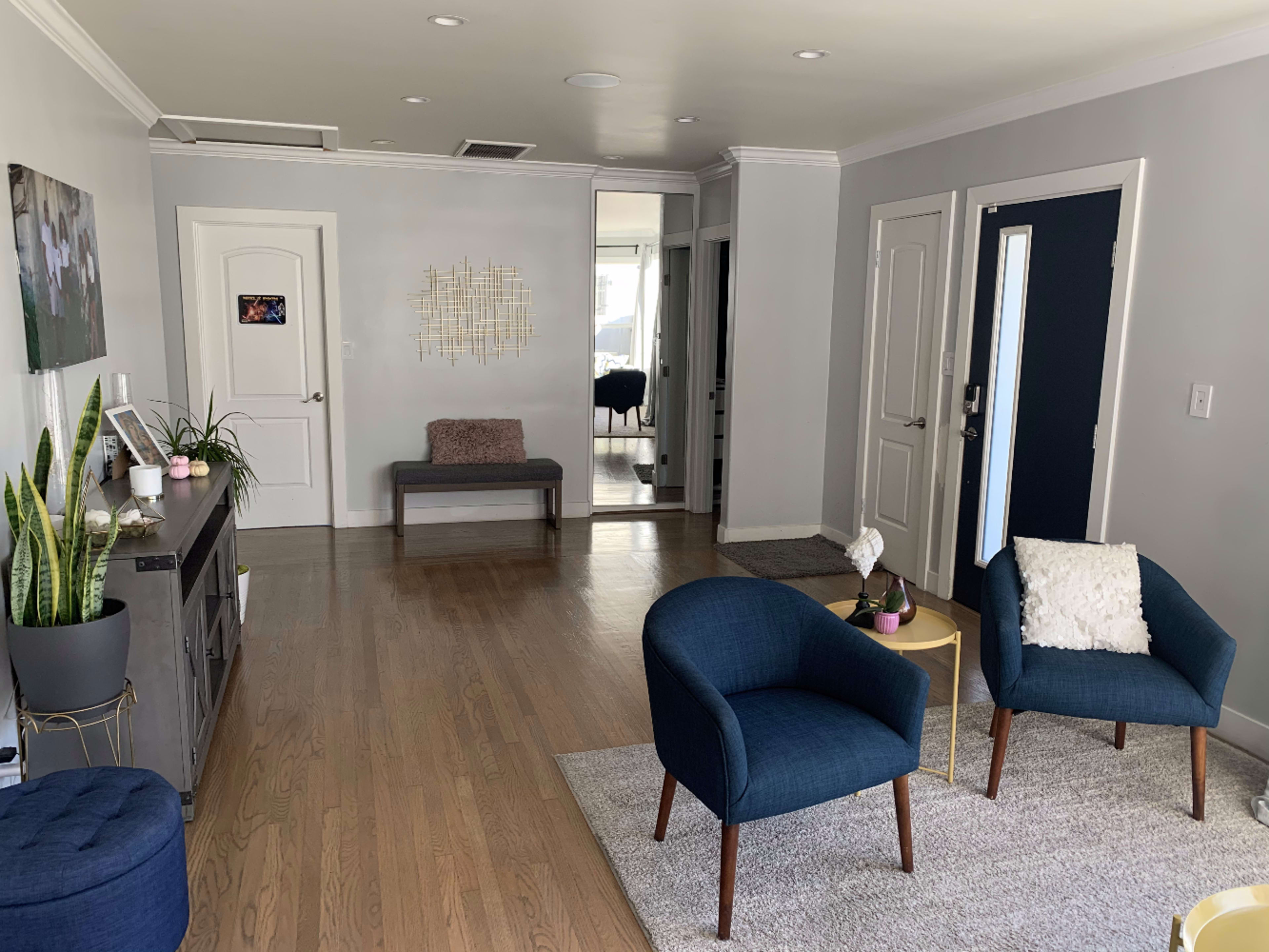 The image shows a modern living space with two blue armchairs, a gray rug, a wooden bench, and a console table against the wall, along with a door and a mirror reflecting light.