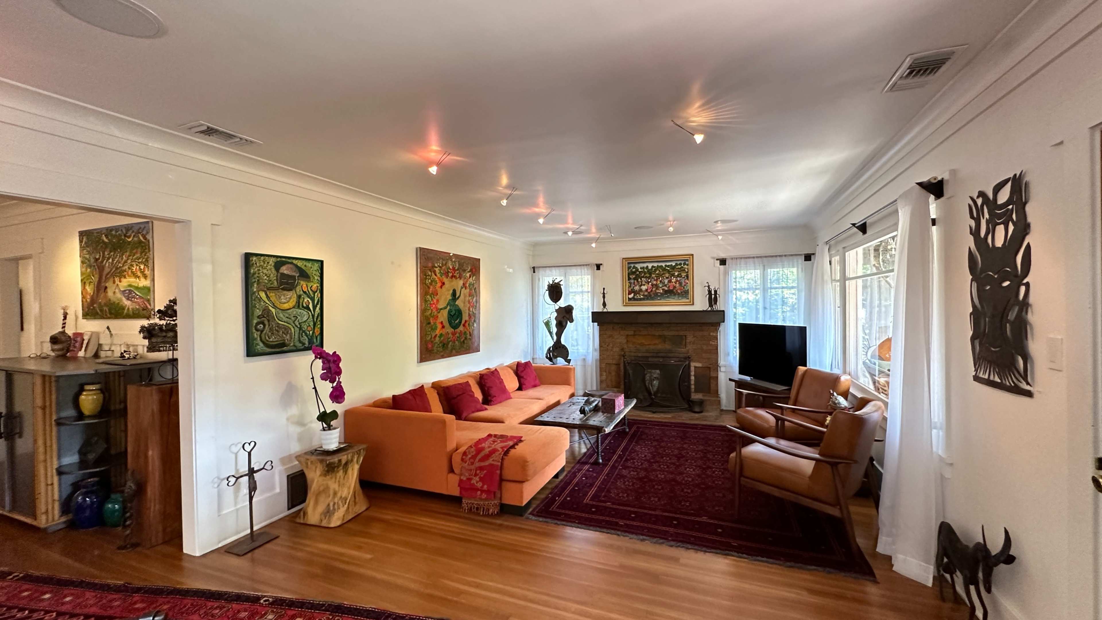 The living room features an orange sectional sofa, two brown leather chairs, a coffee table, and various art pieces on the walls, with a fireplace and large windows allowing natural light.