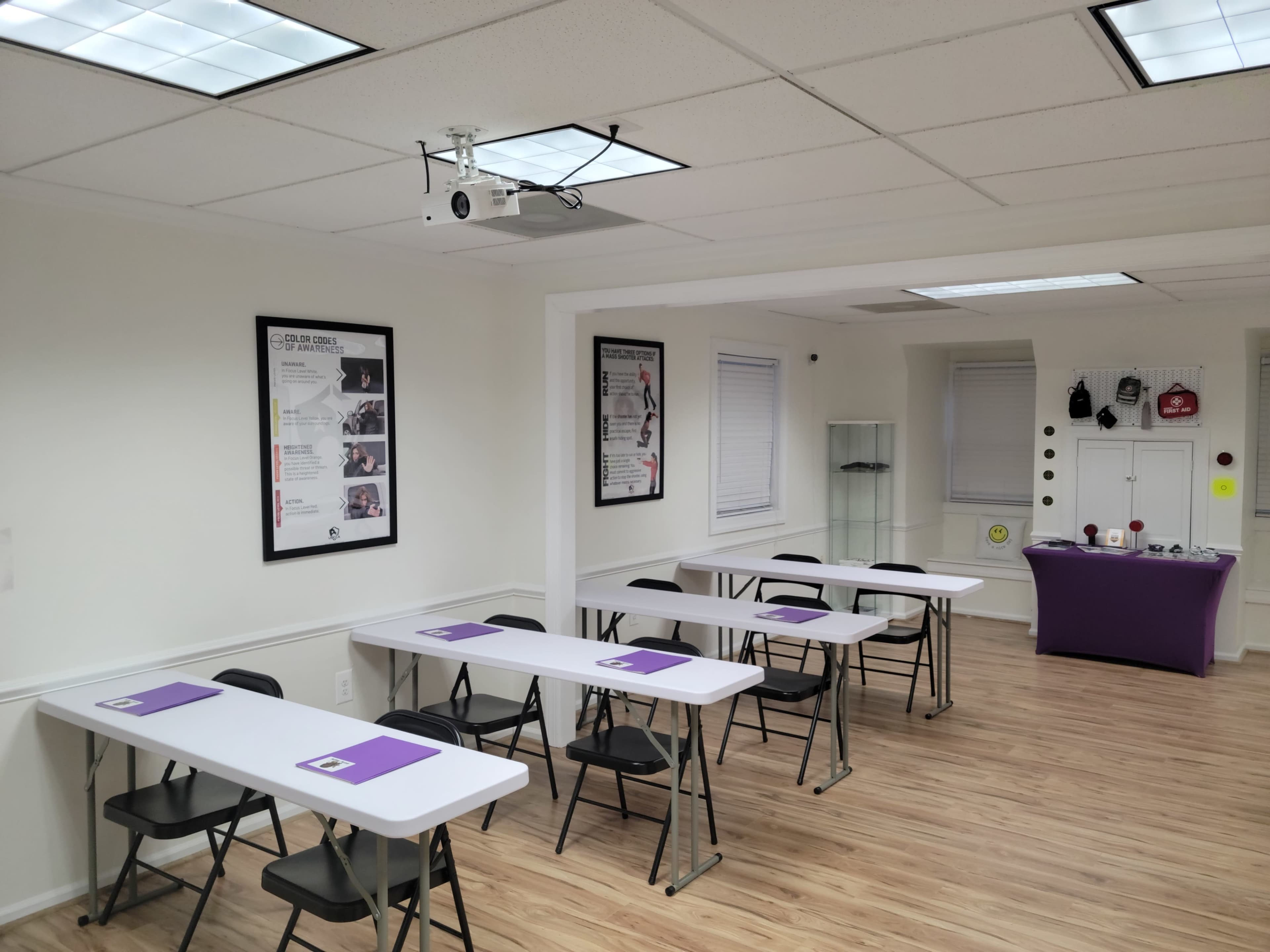 A classroom setting features rows of tables with chairs, a projector mounted on the ceiling, and various instructional materials displayed on the walls.
