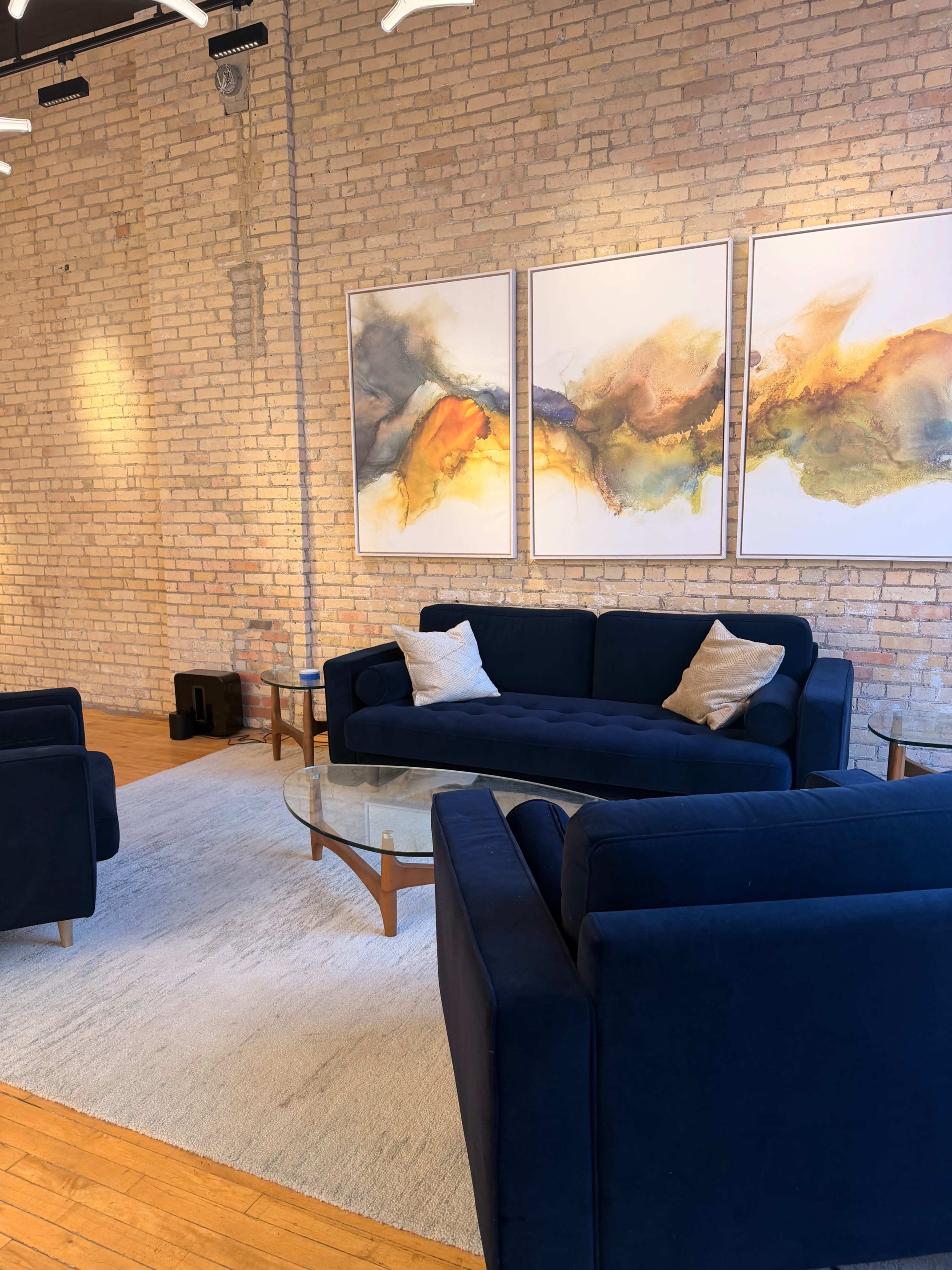 The image shows a modern living room with a blue sofa and armchairs arranged around a glass coffee table on a light-colored rug, featuring three abstract paintings on the brick wall.