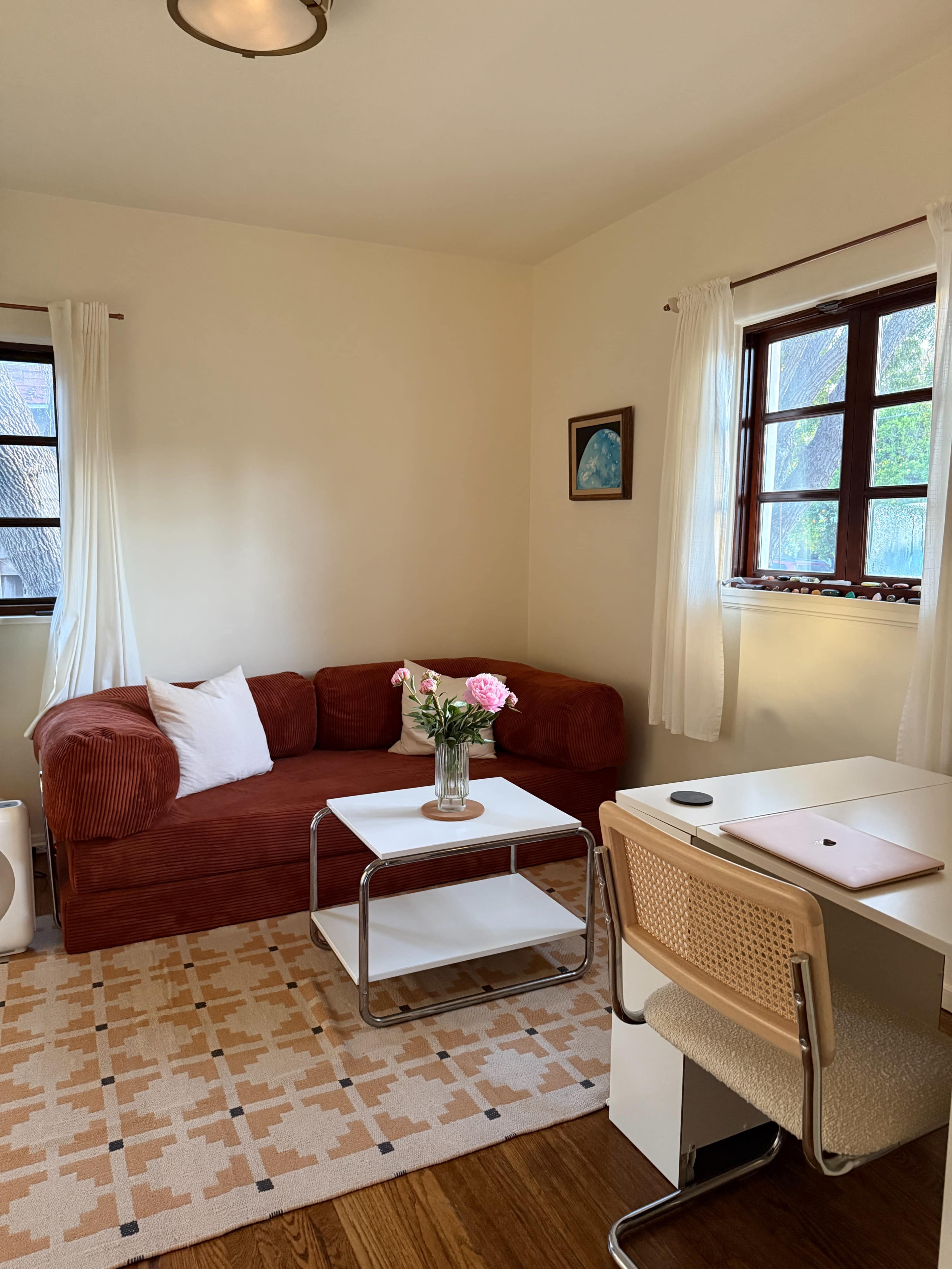 The image shows a small living space with a red couch, a white coffee table, a desk with a chair, and natural light coming through two windows.