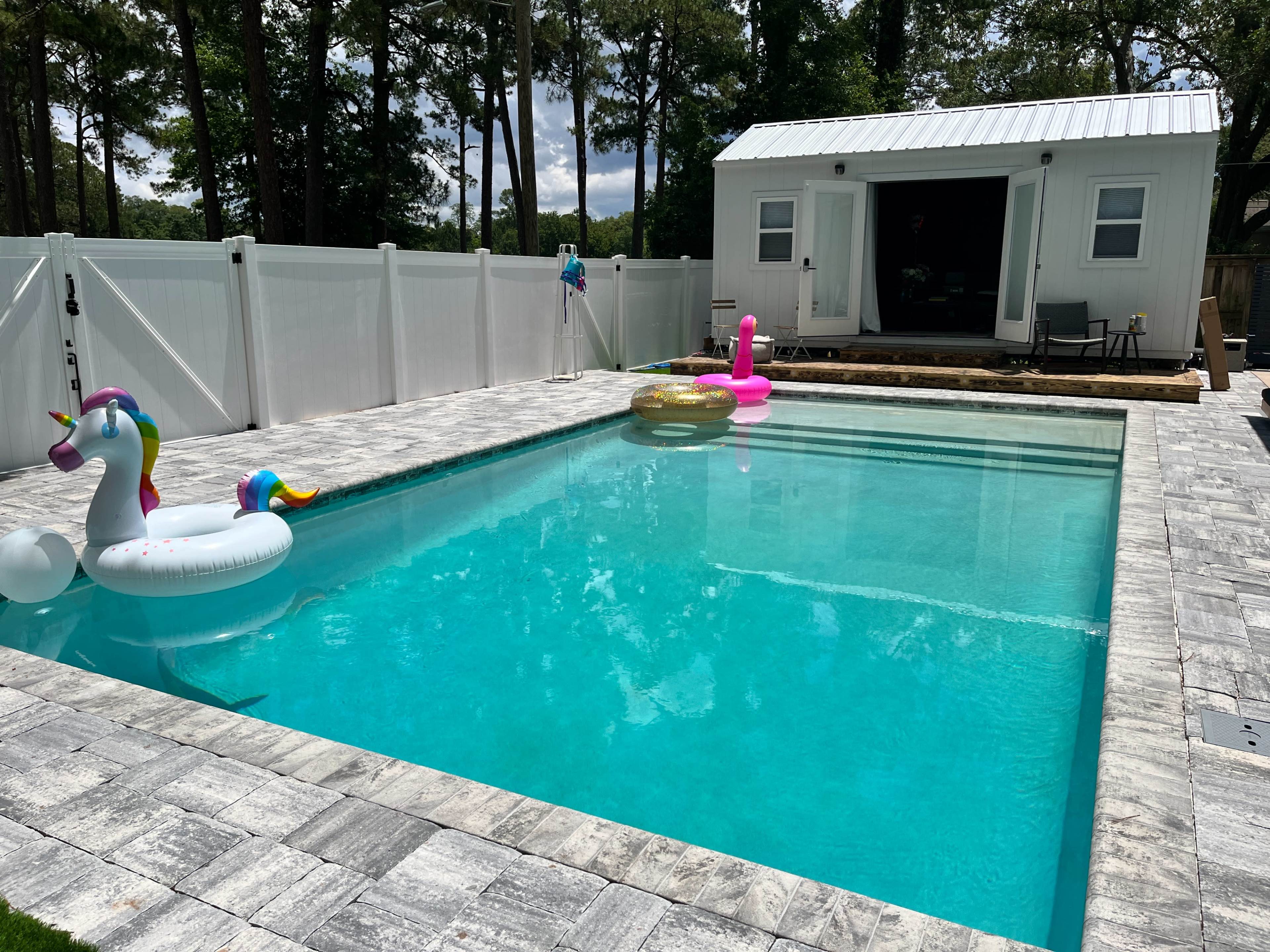 A residential backyard with a clear pool, bordered by a white fence, and two inflatable pool floats, a unicorn and a ring, next to a small shed at the edge of the patio.