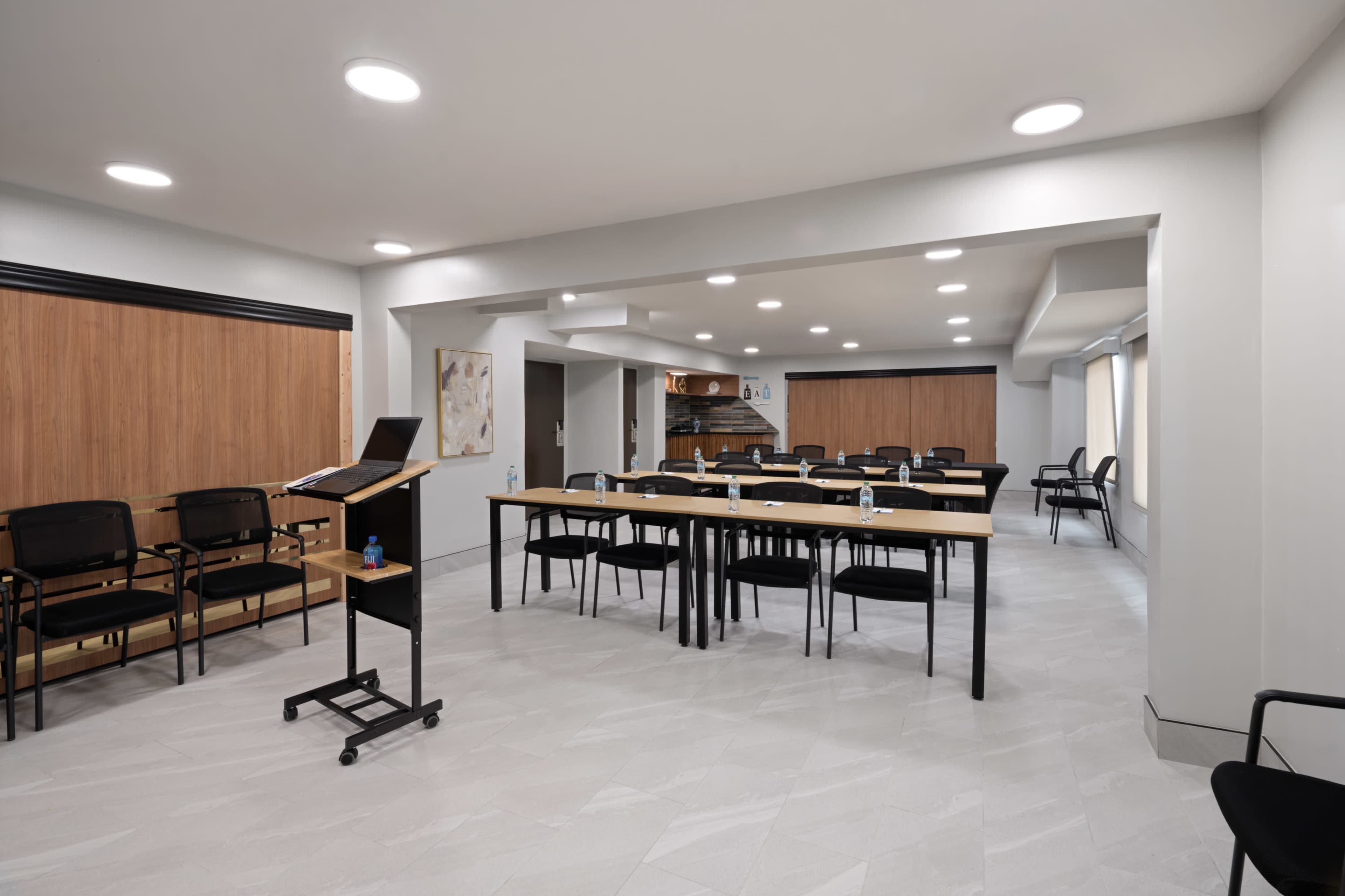 The image shows a well-lit meeting room with several tables and chairs arranged for gatherings, featuring a presentation area at the front.