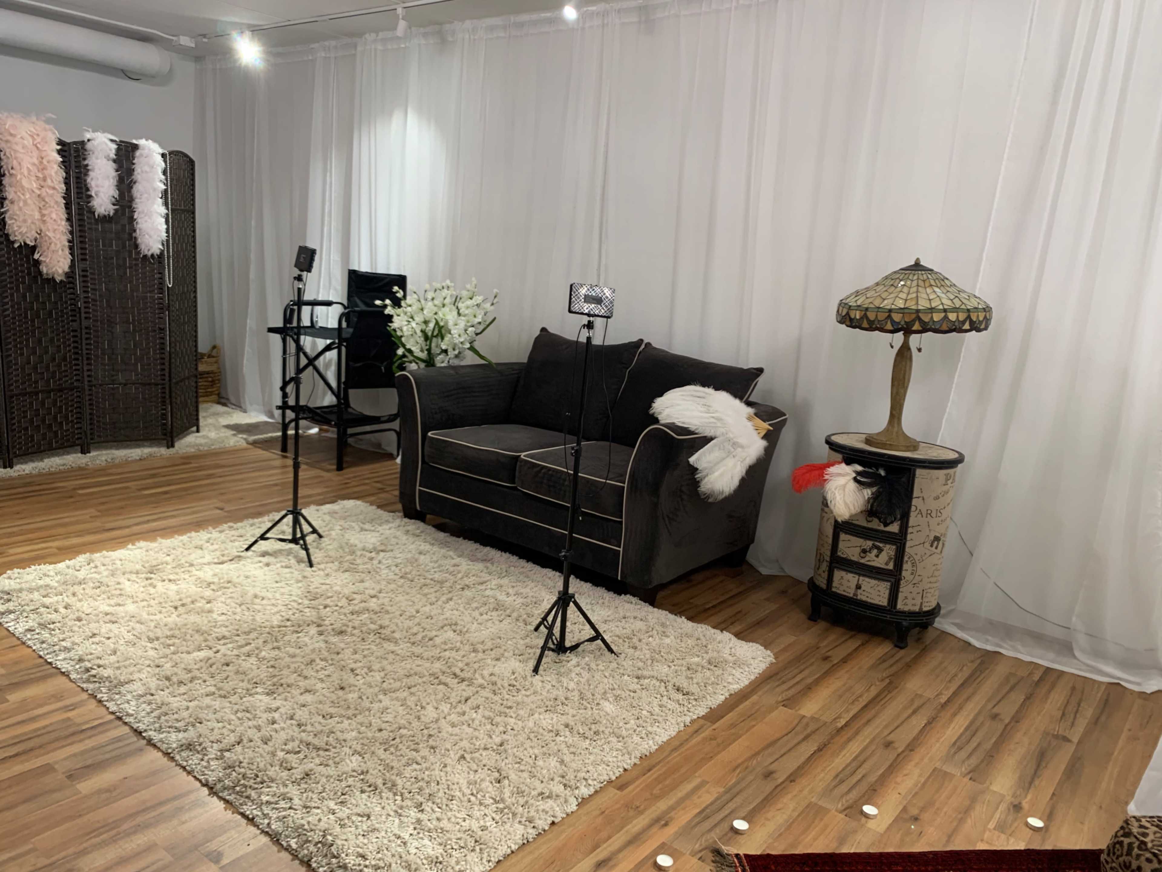 The image shows a cozy studio setup featuring a dark sofa, two lighting stands, a table lamp, and decorative elements on a plush rug.